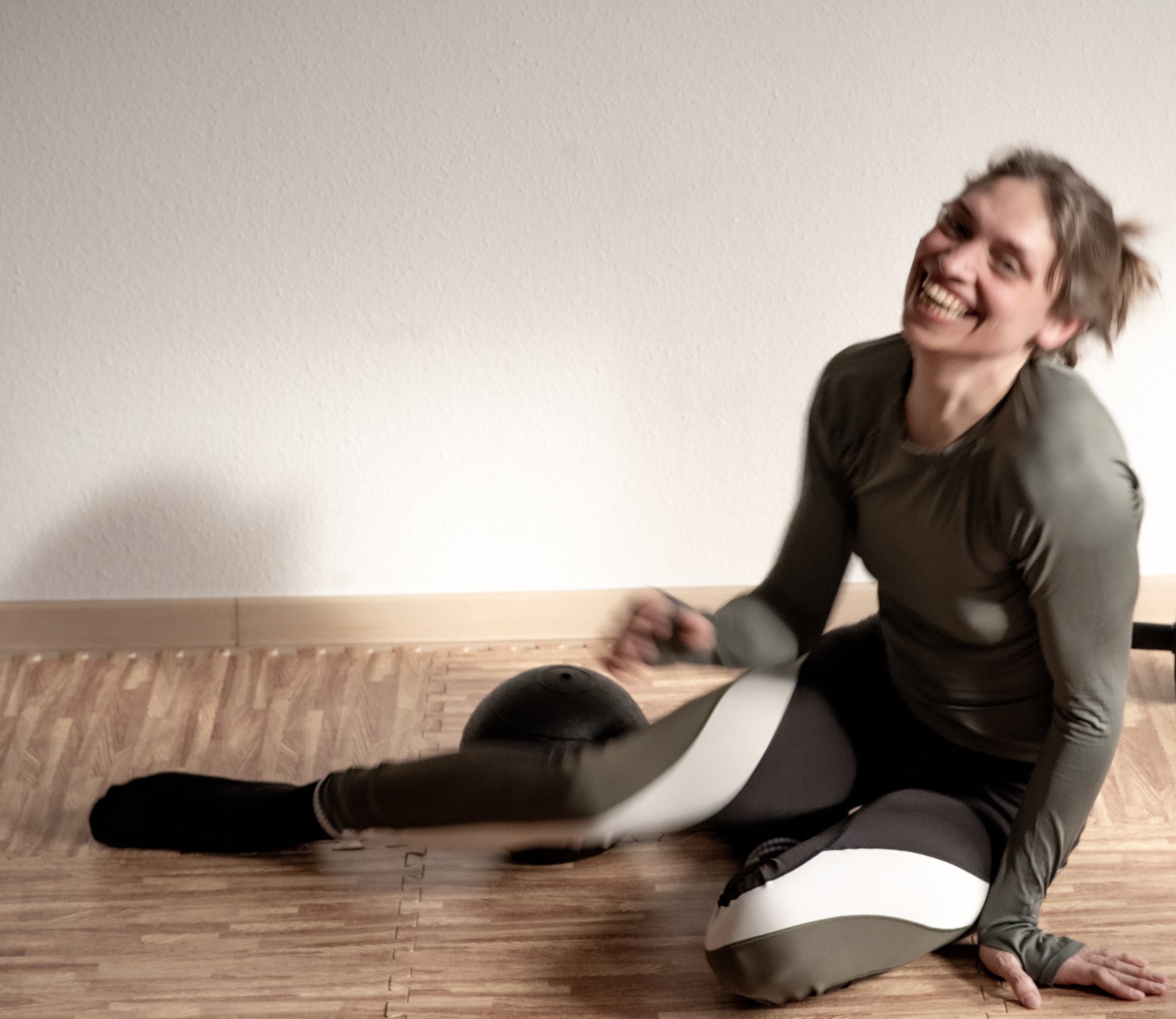 Eine lachende Person trainiert auf dem Boden mit einer schwarzen Medizinball, trägt Sportswear, auf Holzboden vor weißer Wand.