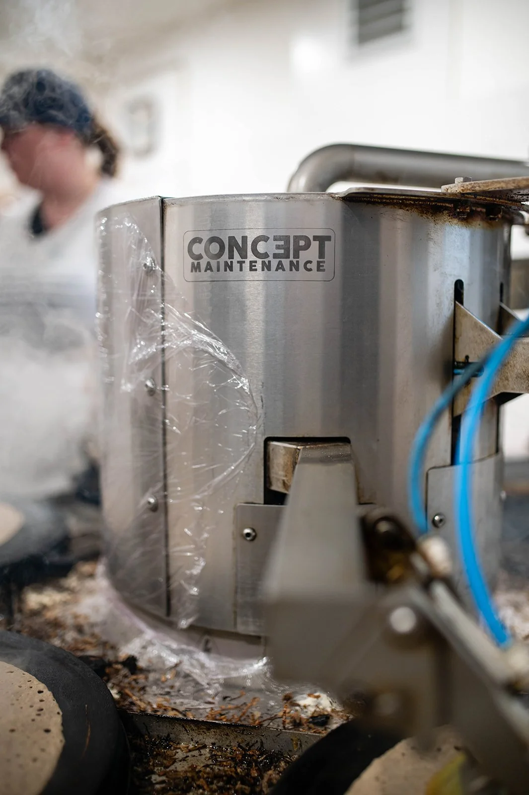 Machine de nettoyage industriel en acier inoxydable portant une étiquette 'CONCEPT MAINTENANCE', dans un environnement de fabrication ou de cuisine, avec quelques débris autour.