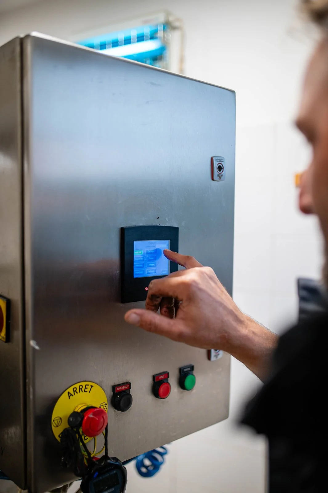 A person using an industrial metal control panel with a touch screen, several buttons, including a red emergency stop button, located in a room with a white wall and a fan in the upper left corner.