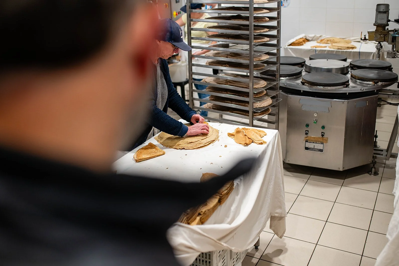 A person is making pancakes in a kitchen, with several pancakes stacked on a table, a pancake rack in the background, and a pancake machine cooking.
