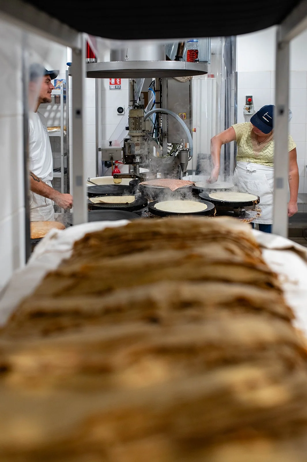 Two people cooking pancakes in a professional kitchen, seen through a metal frame, with stacks of pancakes in the foreground.