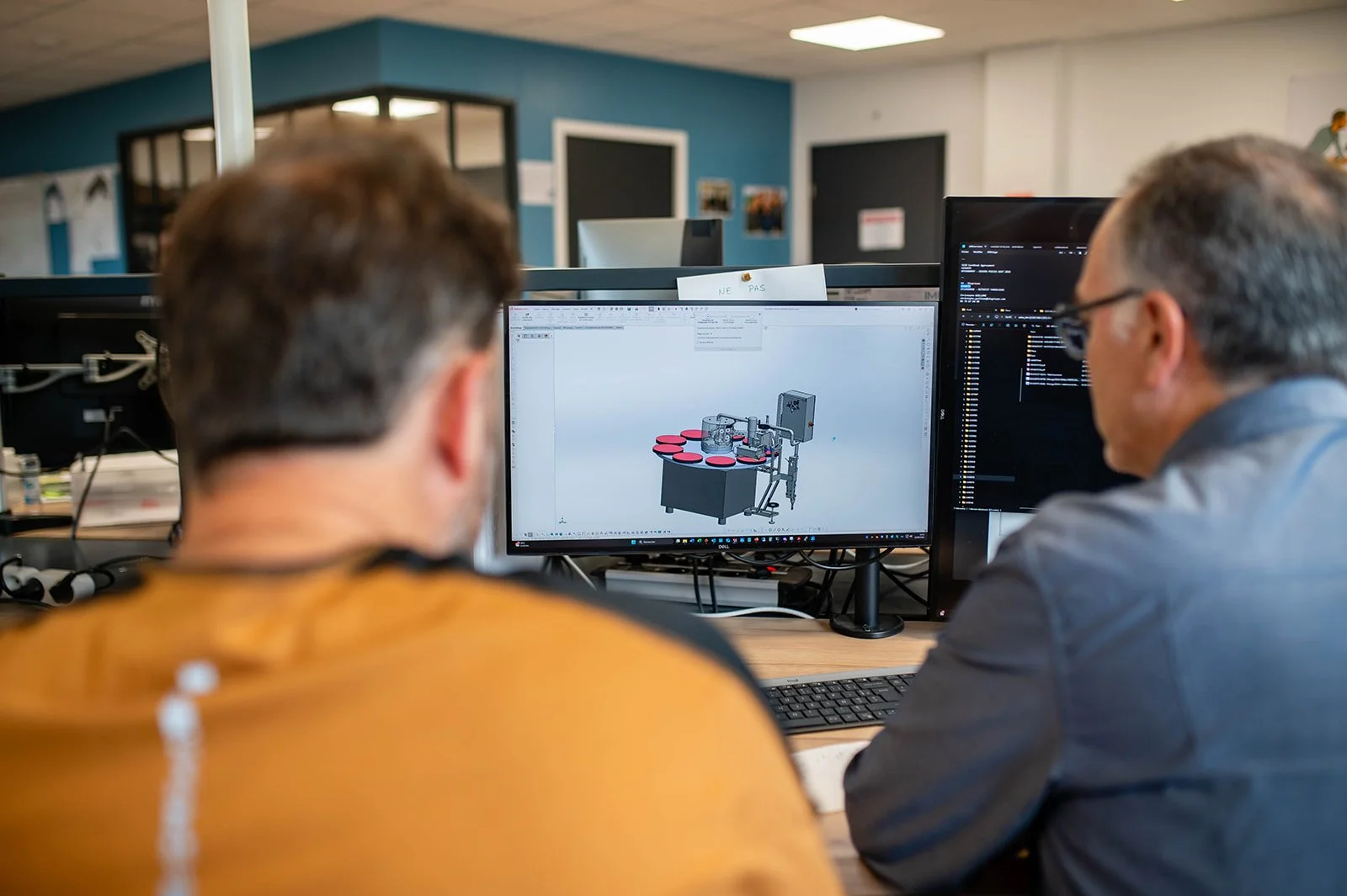 Two men looking at a 3D computer model of a manufacturing or packaging machine on two computer screens in an office.