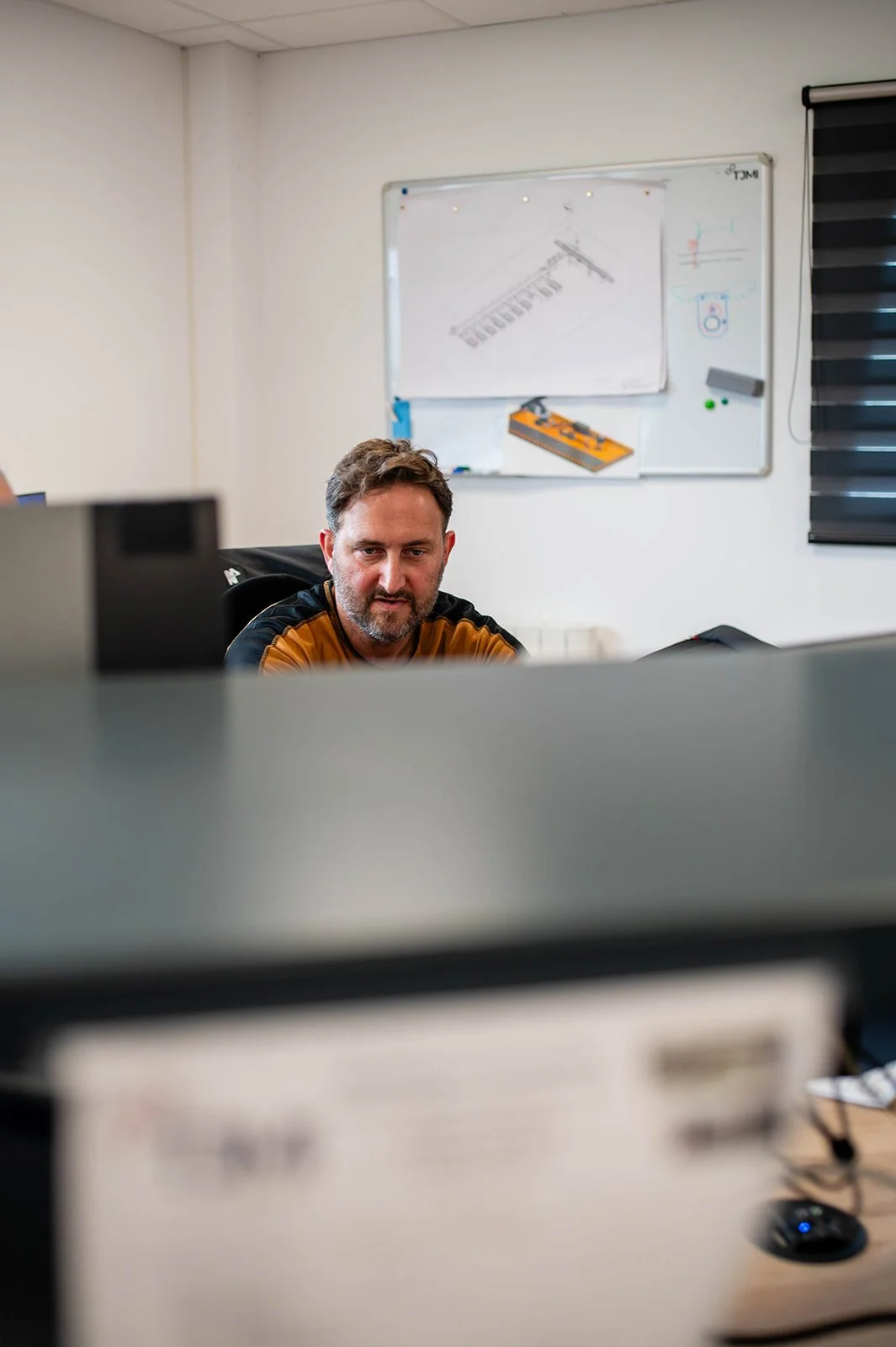 A man sitting at a desk, seen from above a computer, in a room with a whiteboard in the background.