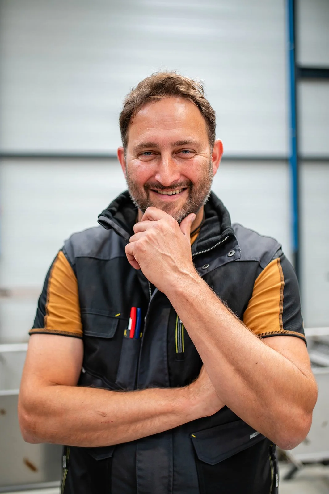 Smiling man with a beard, posing with his hand on his face in an industrial setting.