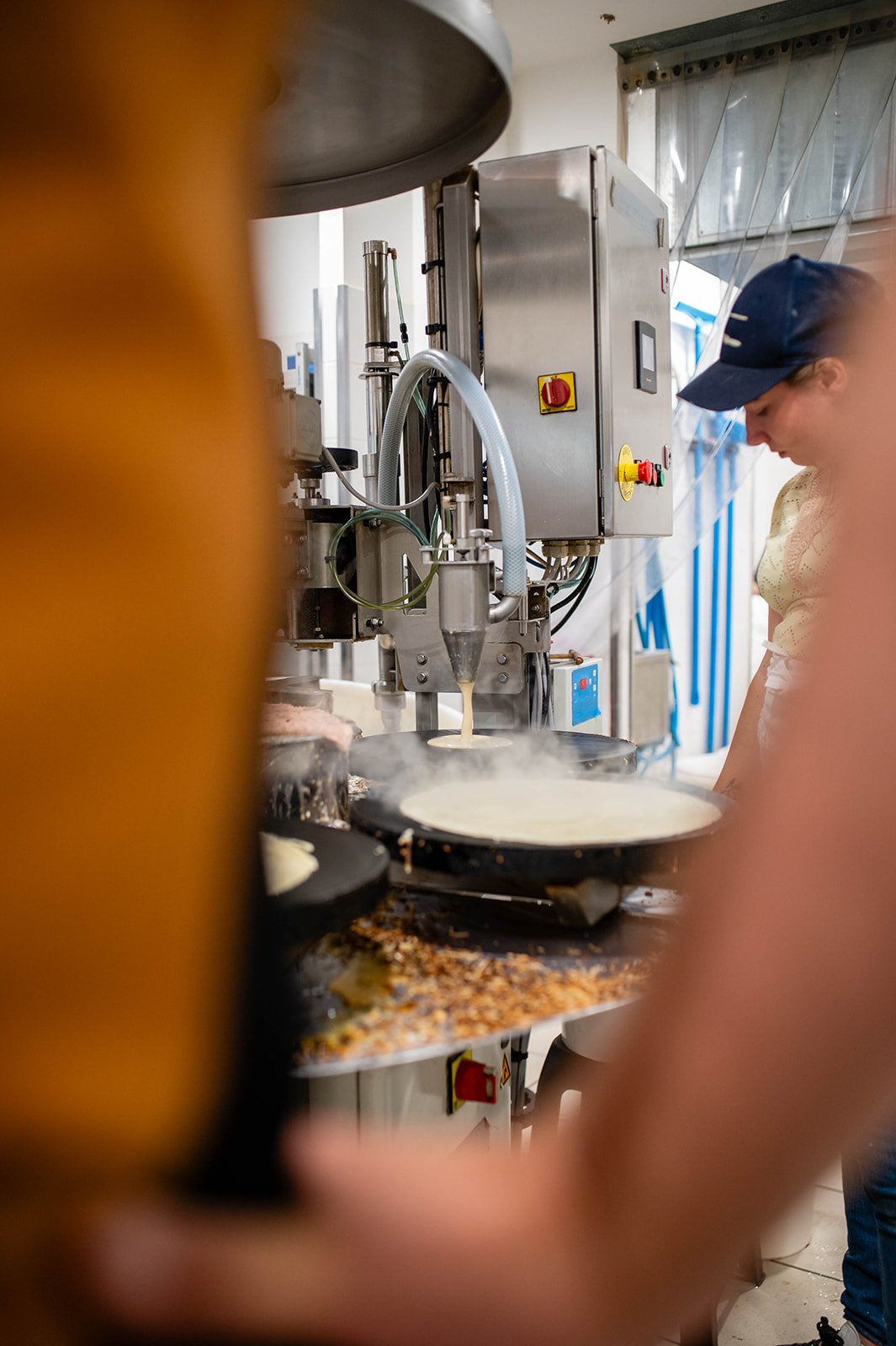 A woman wearing a cap prepares crepes or pancakes with a crepe maker in an industrial kitchen.