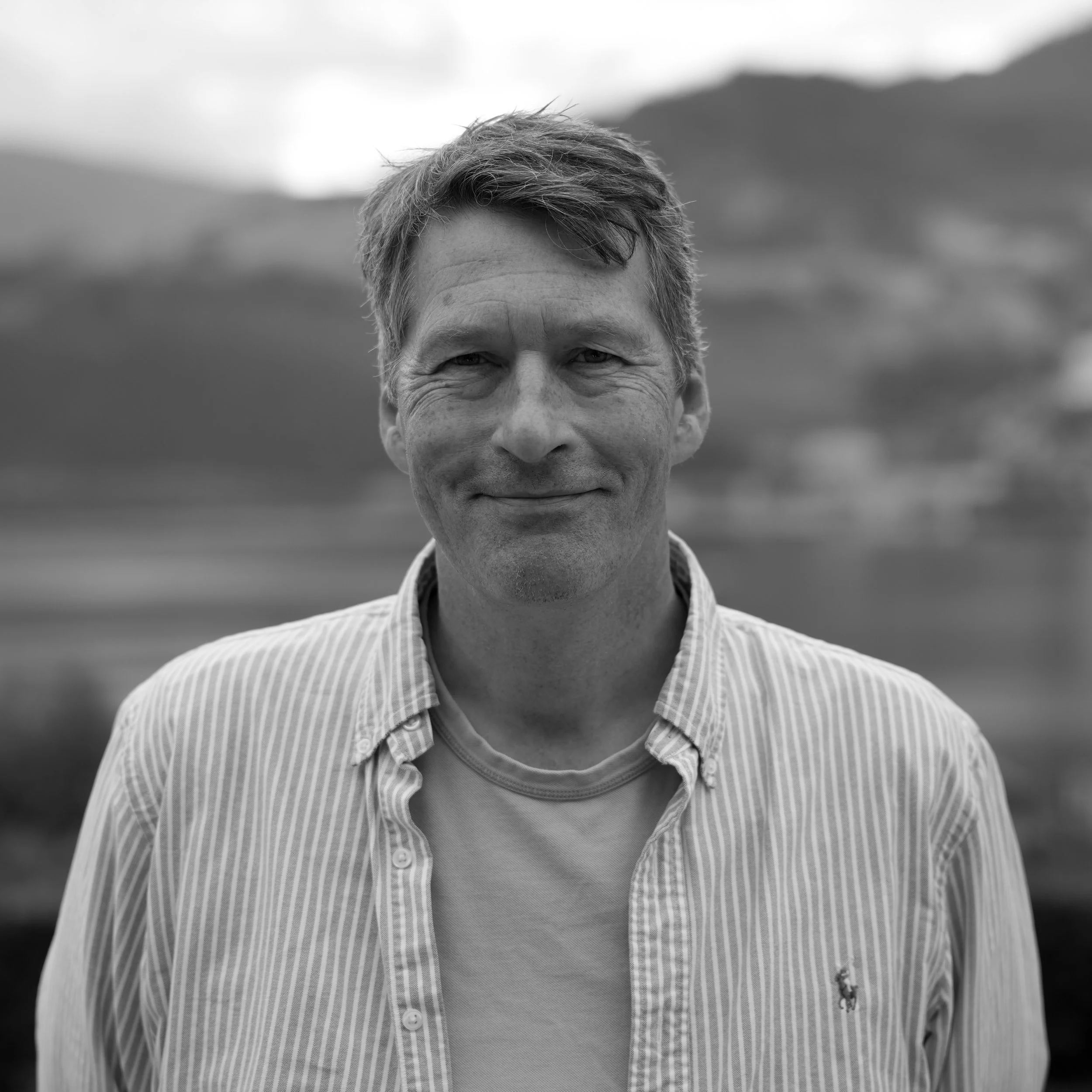 En mann står utendørs foran en bygning med skog i bakgrunnen, smilende mot kamera, i svart-hvitt.