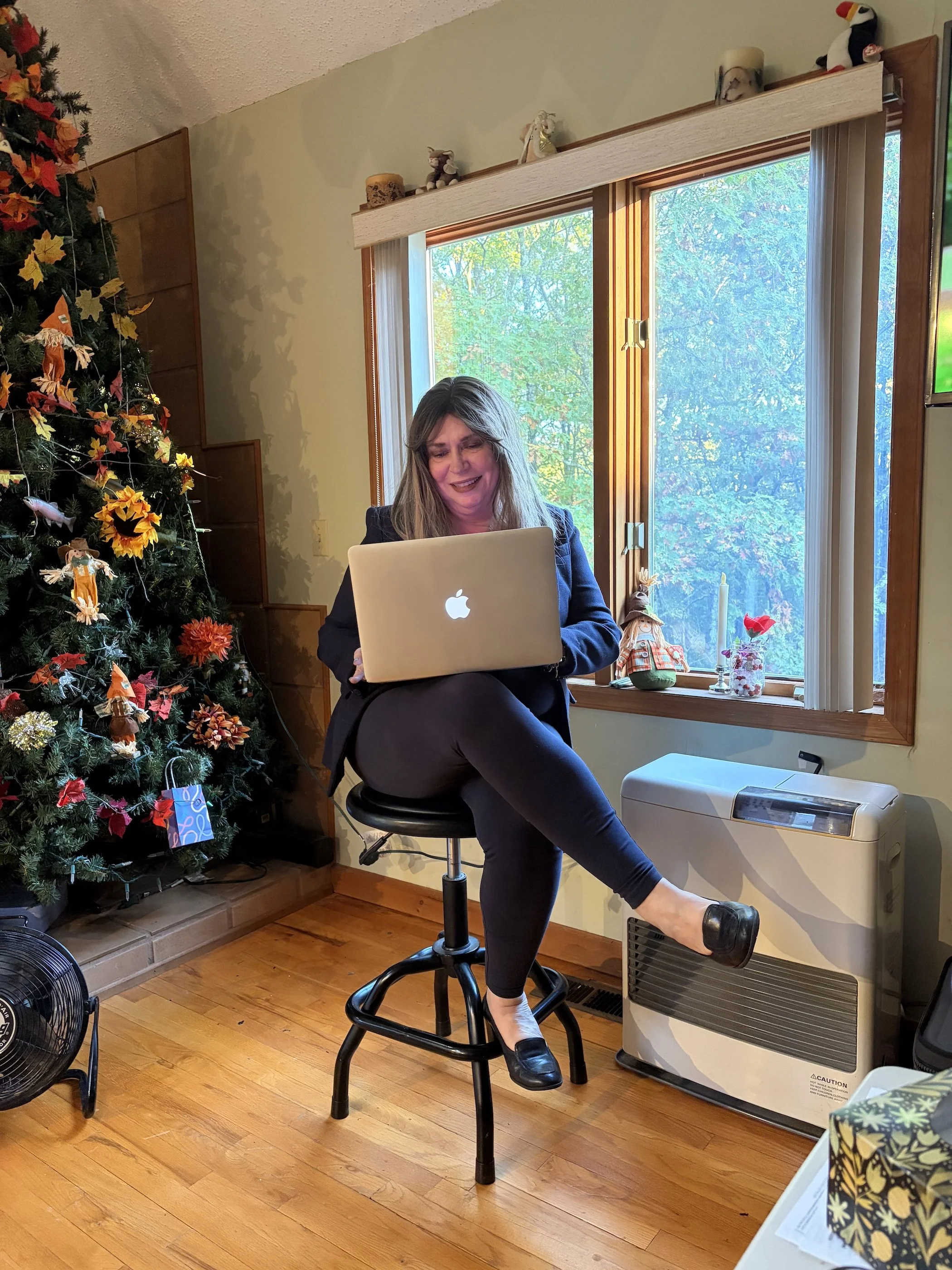 Rita O'Malley sitting on a stool in a room decorated for Christmas, working on a laptop, with a decorated Christmas tree on her left and a window showing trees outside behind her.