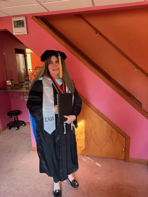 Rita O'Malley in a graduation gown and cap standing indoors next to a staircase with pink walls, smiling at the camera.