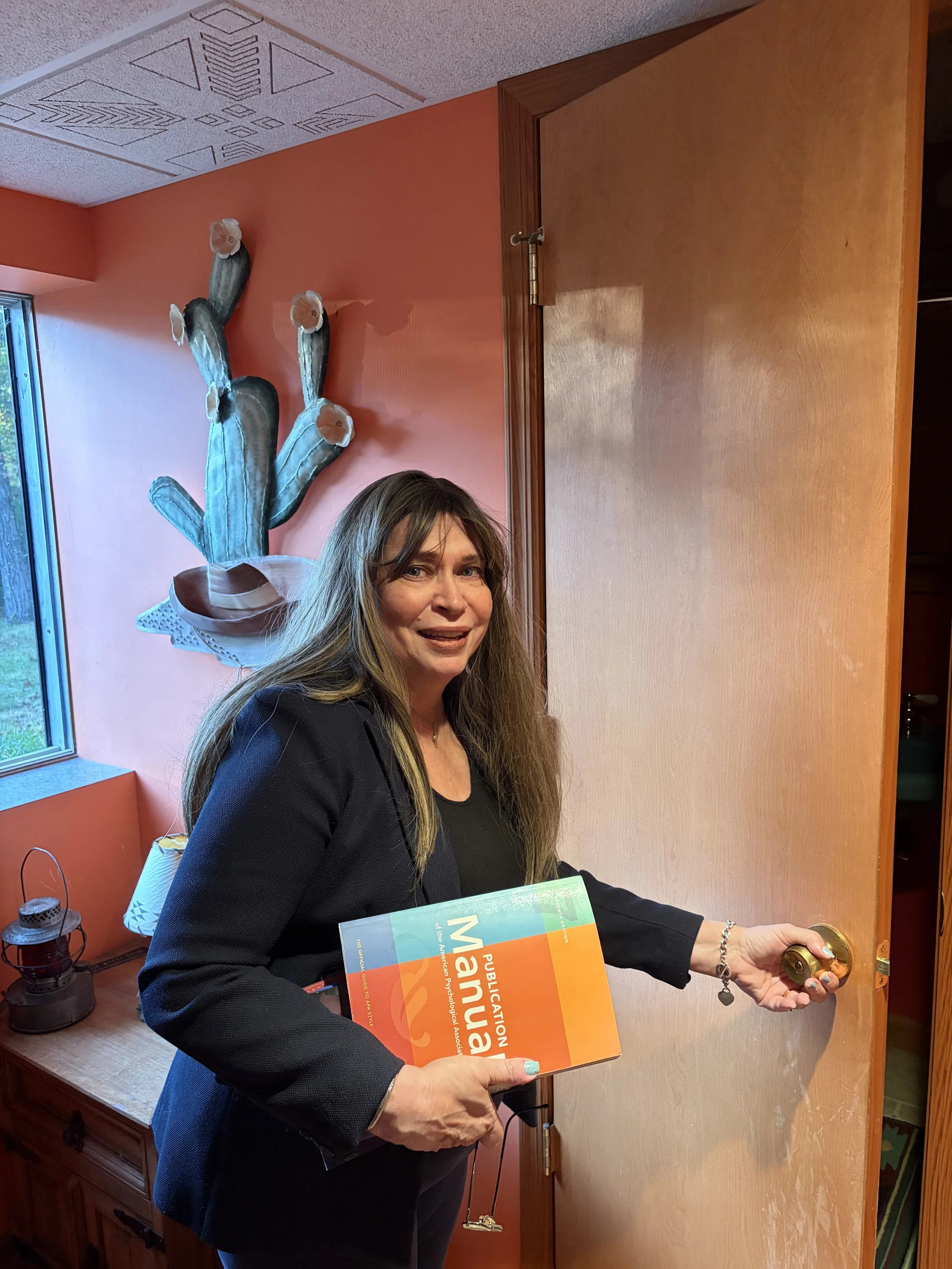 A woman with long hair and a black jacket opening a wooden door while holding a publication and smiling. Behind her is a decorative cactus sculpture on a pink wall, and a window with a lantern on a table nearby.
