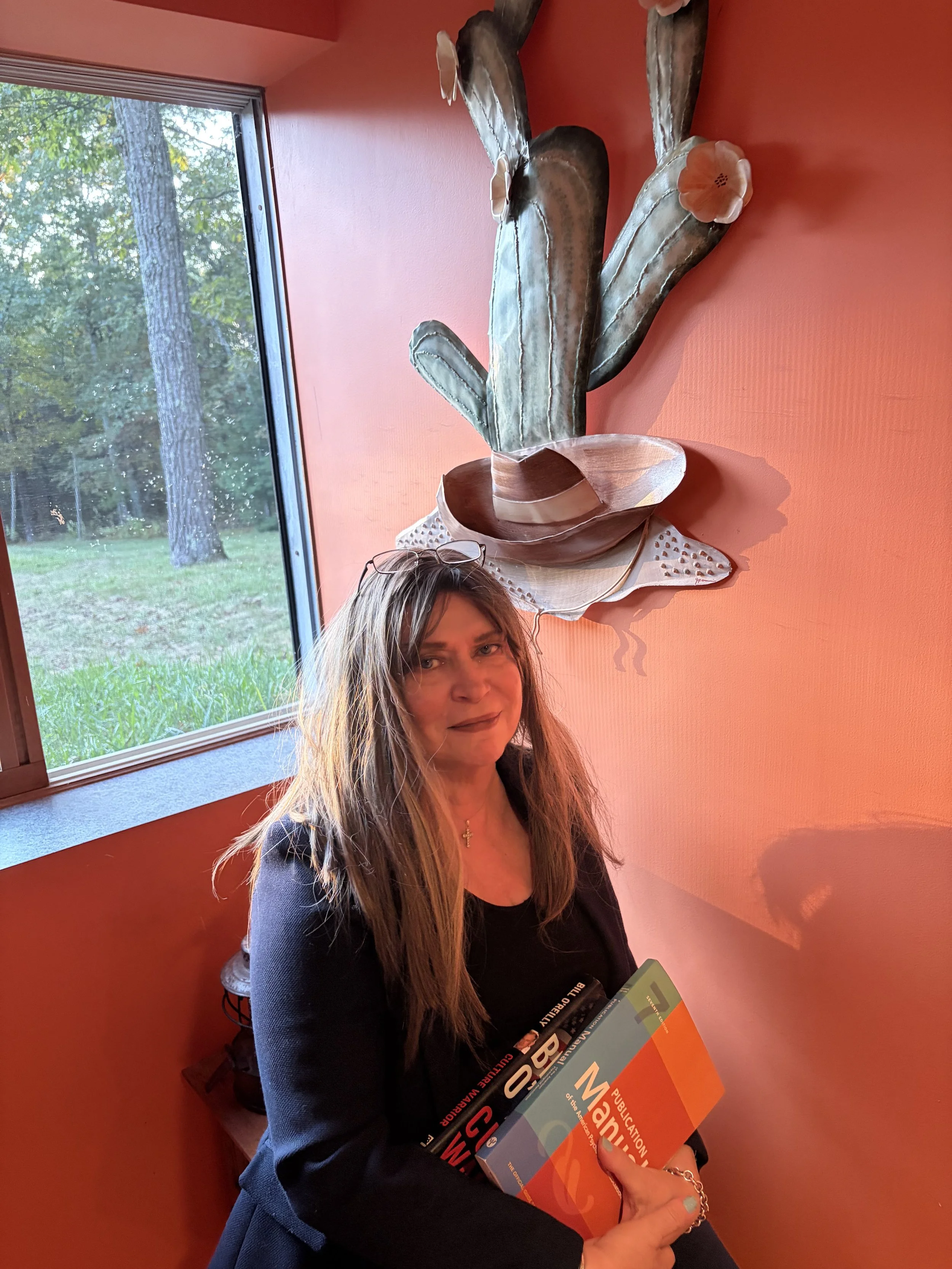 Rita O'Malley holding books and sitting in a room with orange walls and a large cactus wall decoration.