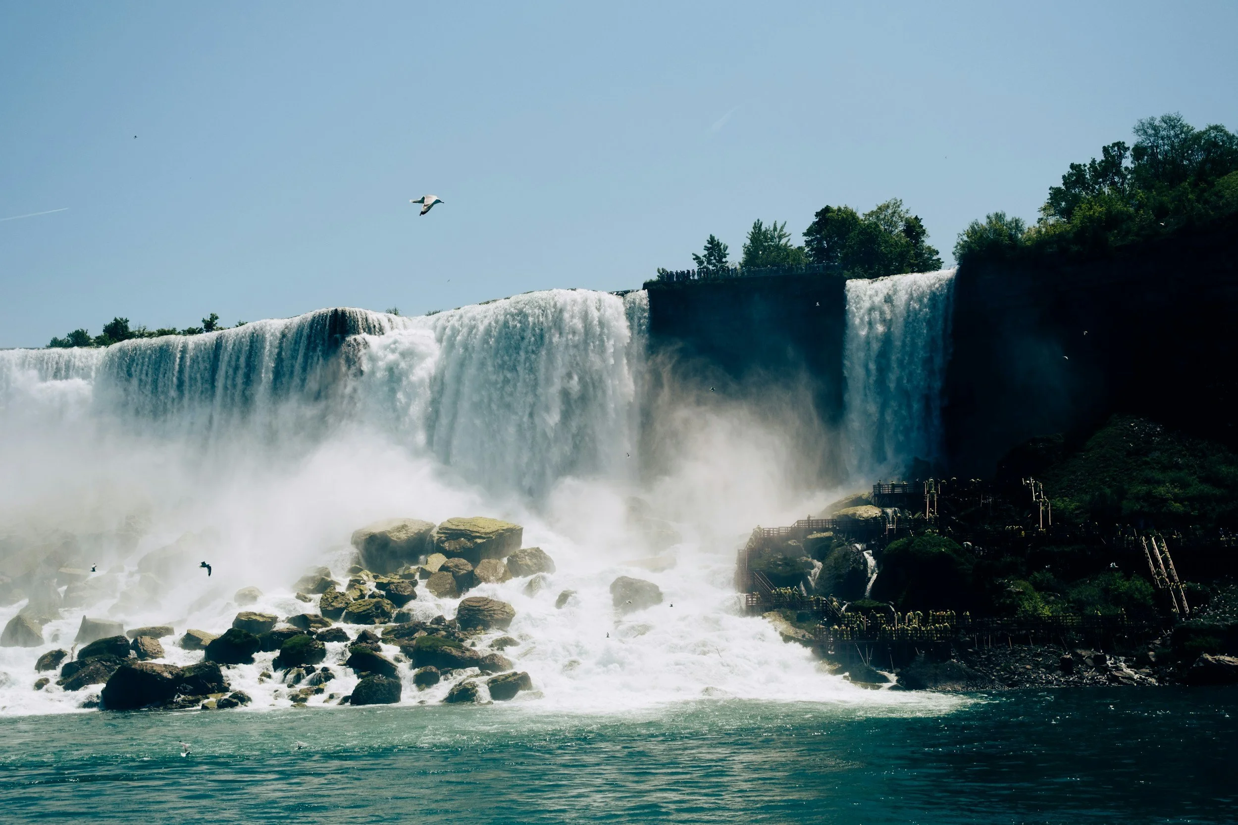 A large waterfall flowing into a river with rocks, surrounded by trees and a clear sky.