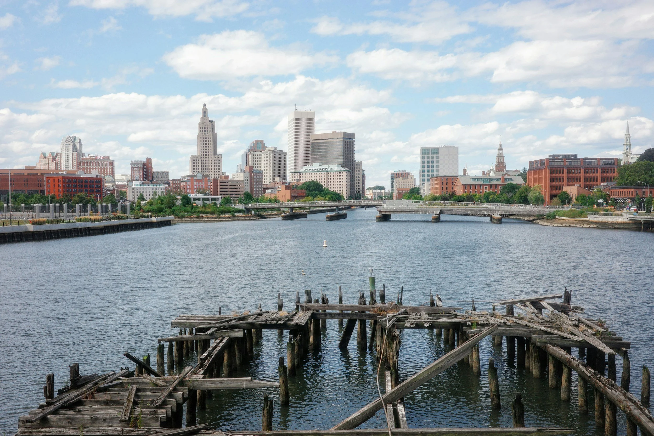 View of a city skyline across a body of water with an old wooden dock in the foreground, under a partly cloudy sky.