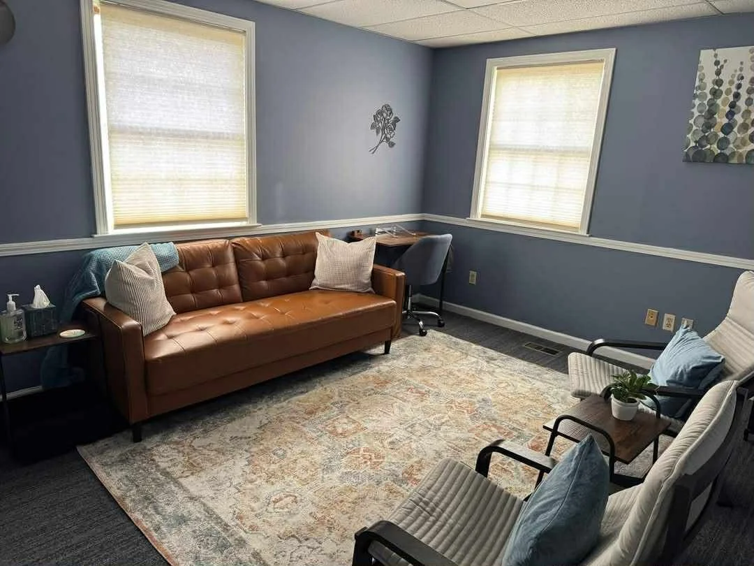 Therapy room with blue walls, brown sofa, gray chairs, small wooden table with a plant, two windows with white blinds, area rug, desk with a chair, and wall decorations.