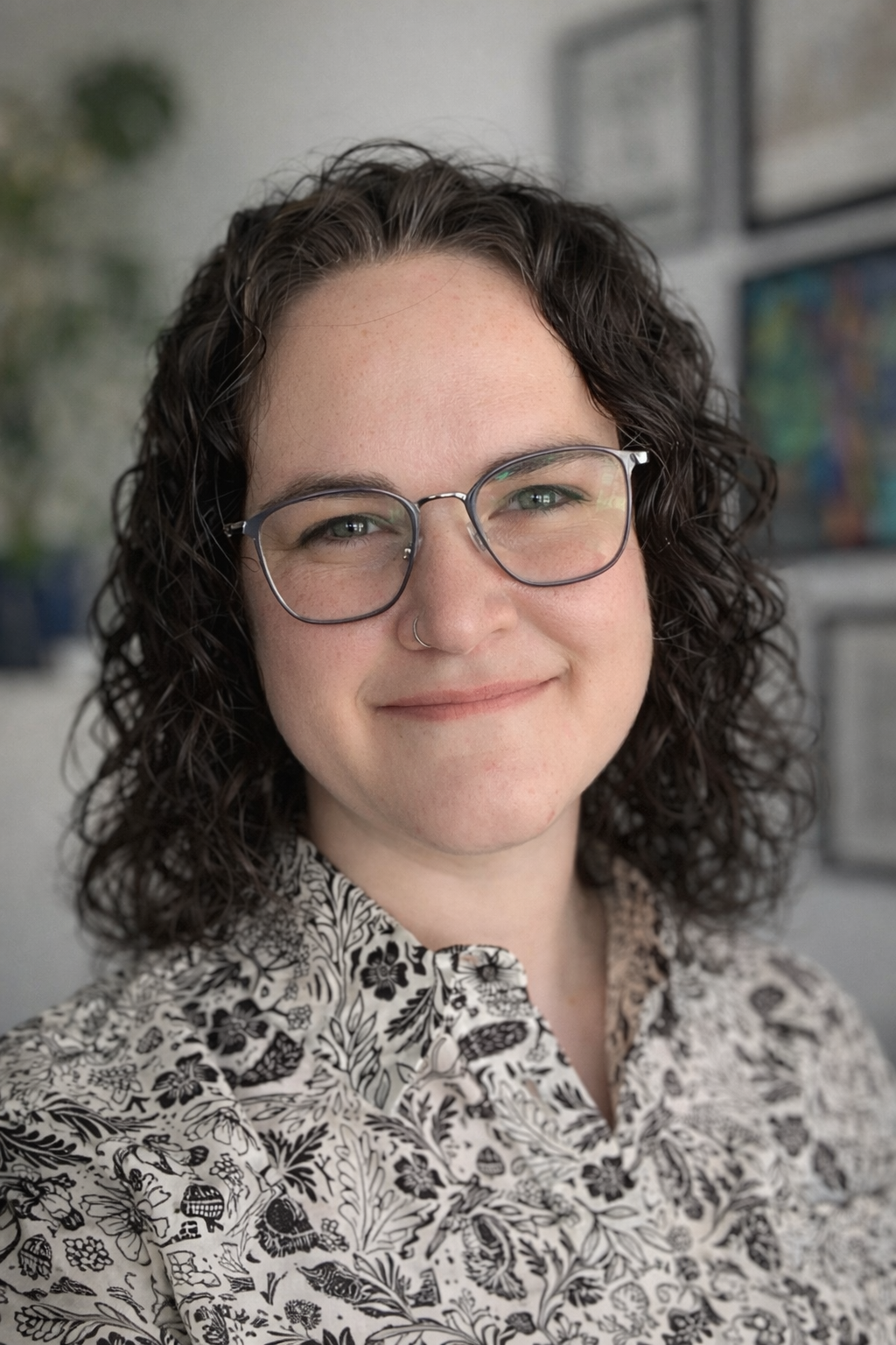 A woman with curly dark hair, glasses, and a nose ring smiling, wearing a black and white floral patterned shirt.
