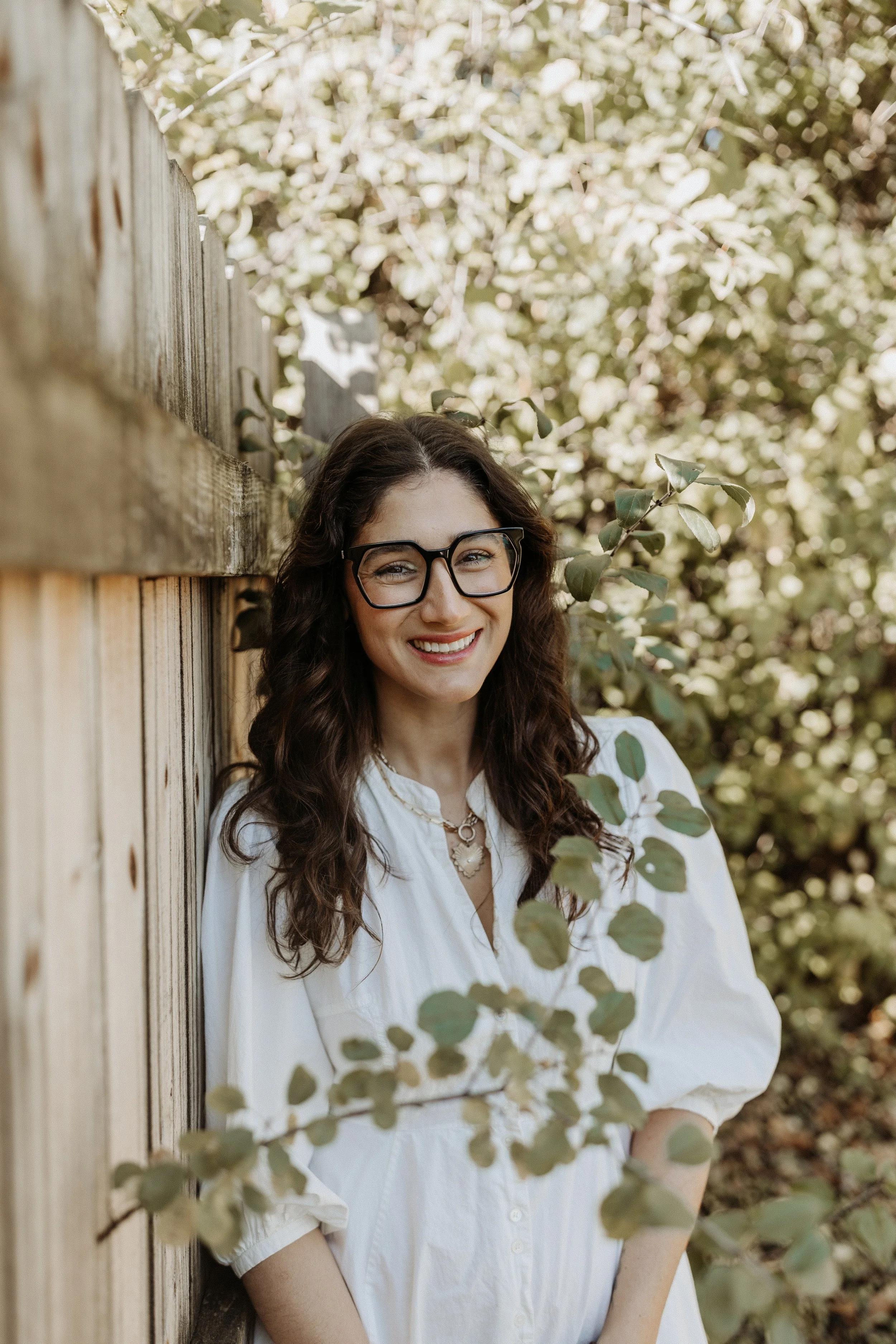 A woman with dark curly hair and glasses smiling at the camera, standing outdoors next to a wooden fence, surrounded by green foliage.