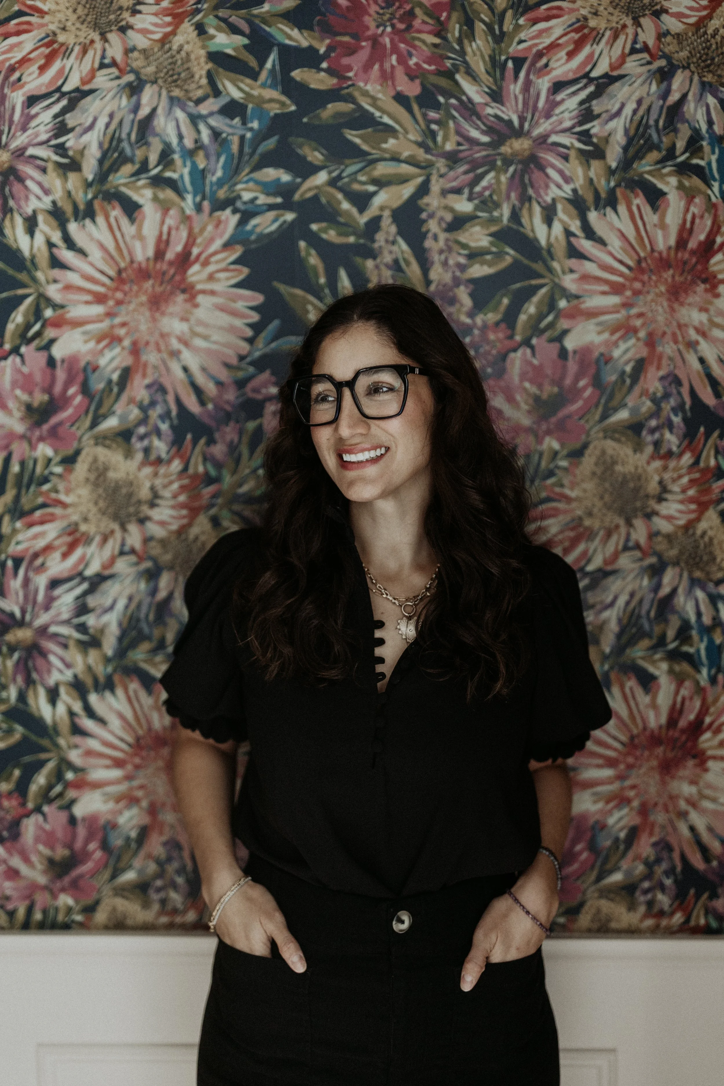 A woman with long curly dark hair and glasses, wearing a black blouse and standing with her hands in her pockets, smiling in front of a floral wallpaper background.
