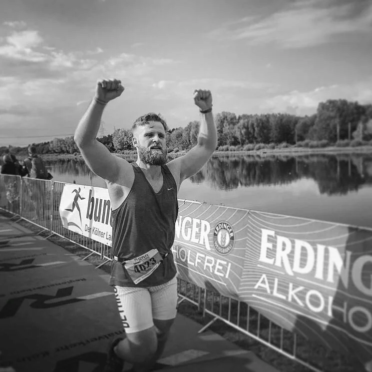 Mann, der bei einem Lauf-Event auf einer Straße an einem Fluss teilnimmt, jubelt mit erhobenen Armen, im Hintergrund Bändchen mit Sponsoren und ein ruhiger Fluss mit Bäumen auf der anderen Seite. Das Bild ist in Schwarz-Weiß.