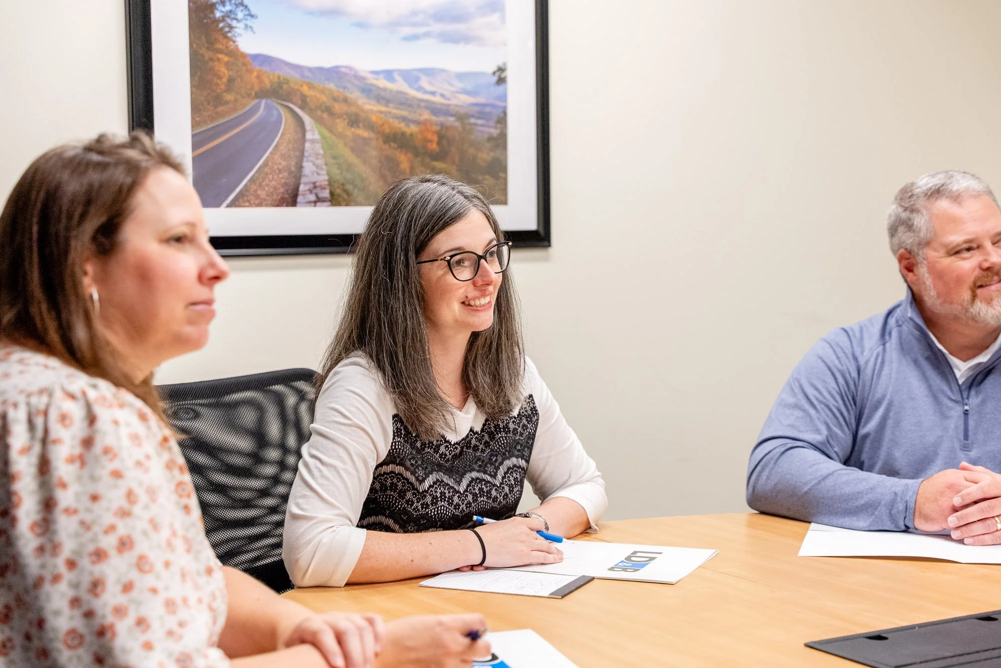 Three people sitting at a conference table in a meeting room, smiling and taking notes, with a framed landscape photograph on the wall behind them.
