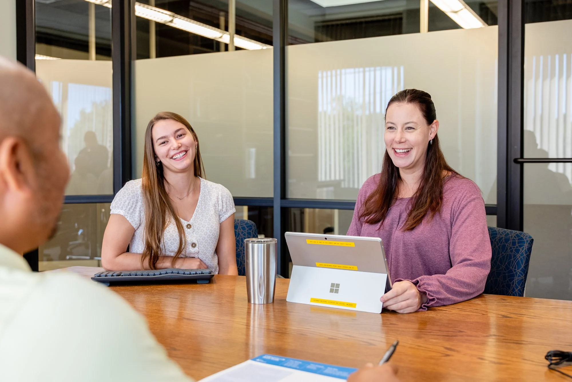 Three women sitting at a conference table, smiling and engaging in conversation. Two women are clearly visible, one on the left with long brown hair wearing a white blouse, and one on the right with dark brown hair wearing a mauve top with a laptop in front of her. A man with a bald head and light shirt is partially visible on the left side. The background shows glass walls with reflections and a modern office environment.