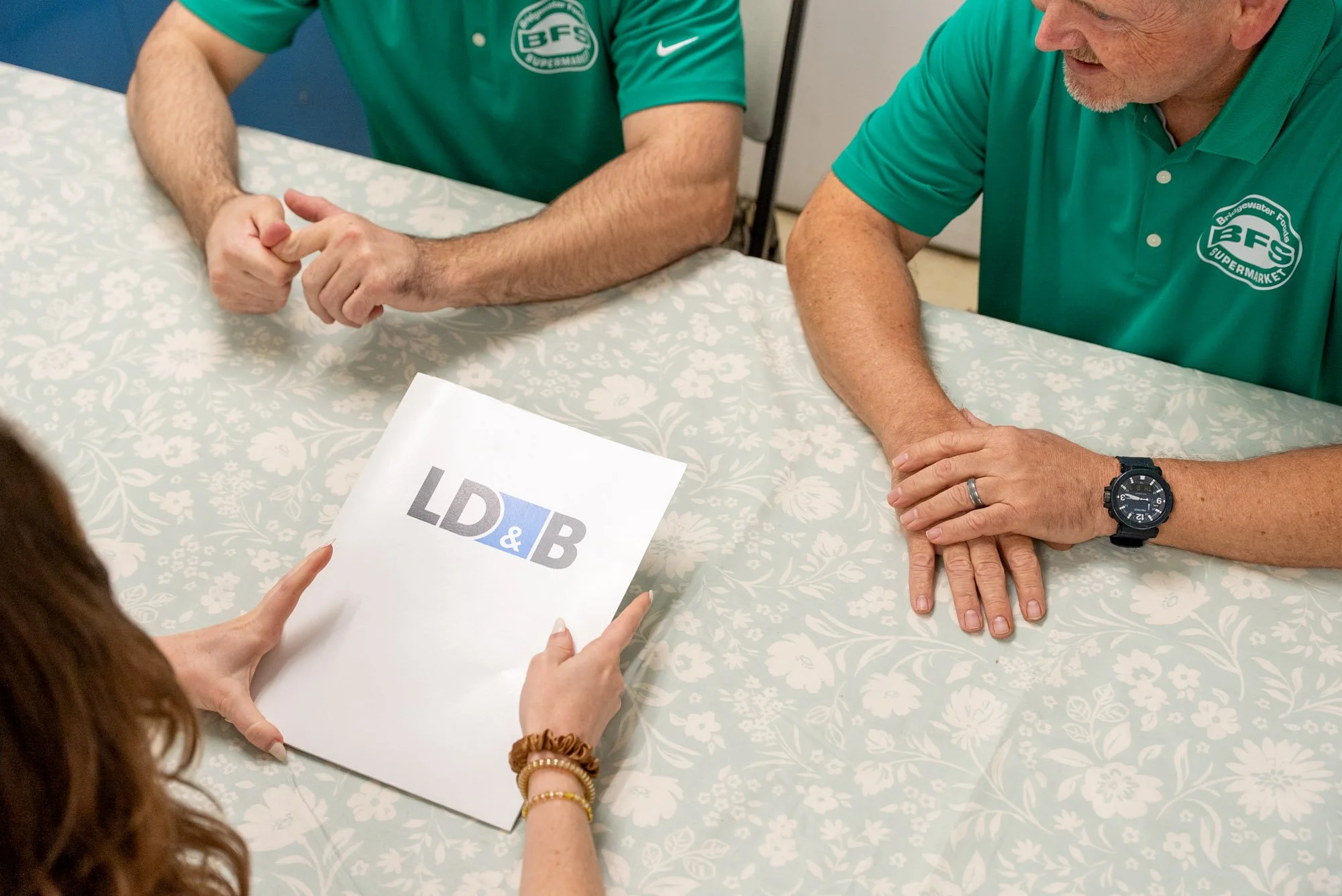 Three people sitting at a table during a meeting or discussion, with a woman holding a LD&B folder.