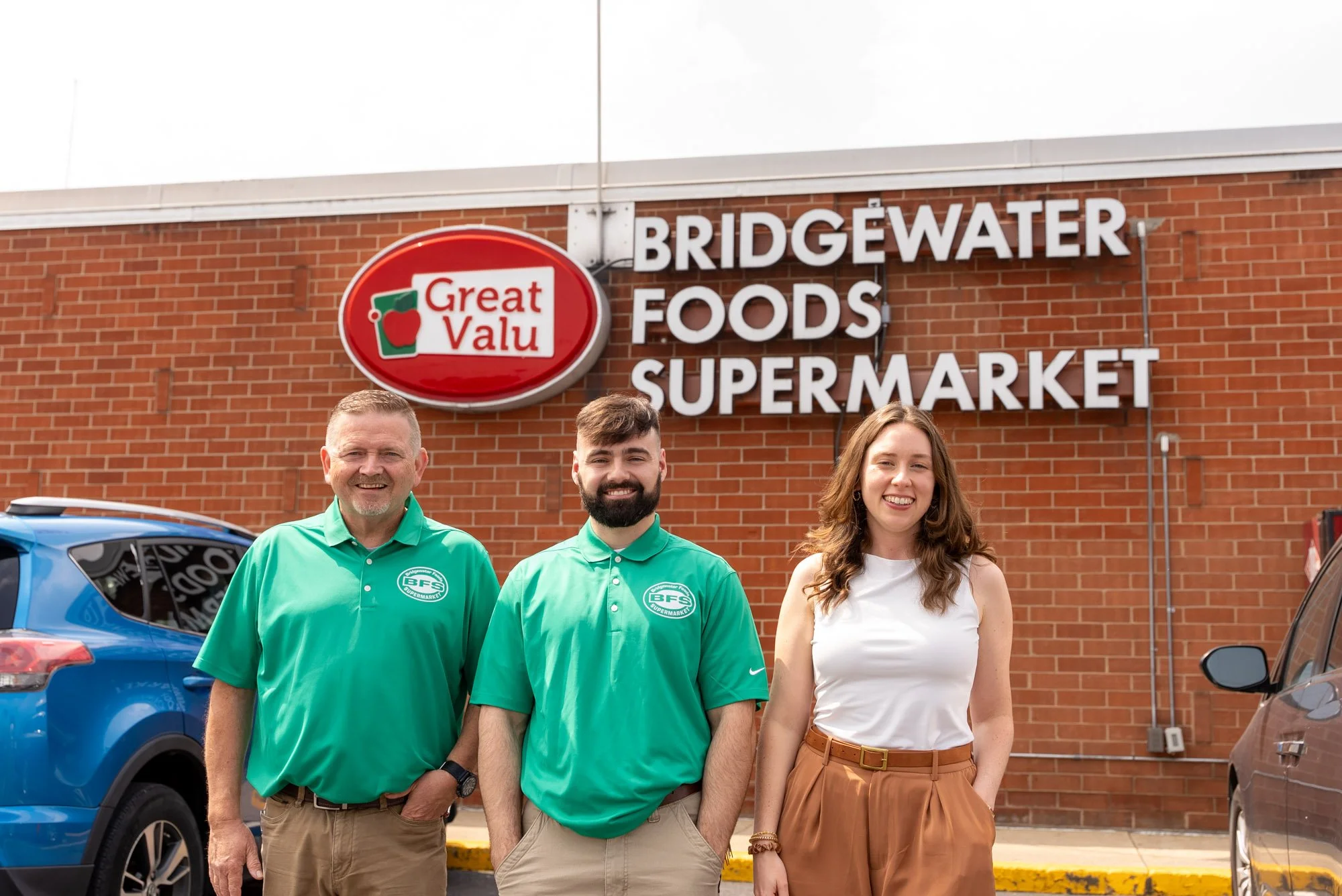 Three people standing in front of a brick store with a sign that reads 'Bridgewater Foods Supermarket' and a logo that says 'Great Valu'. The man on the left and the man in the middle are wearing green polo shirts. The woman on the right is wearing a white sleeveless top and brown pants.