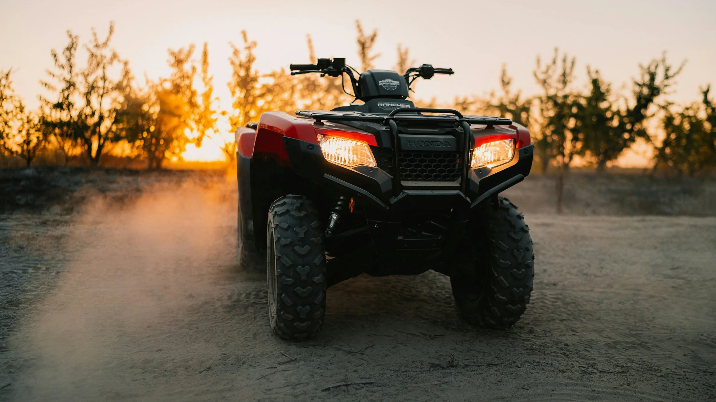 Black and red all-terrain vehicle on dusty dirt ground with trees and sunset in the background.