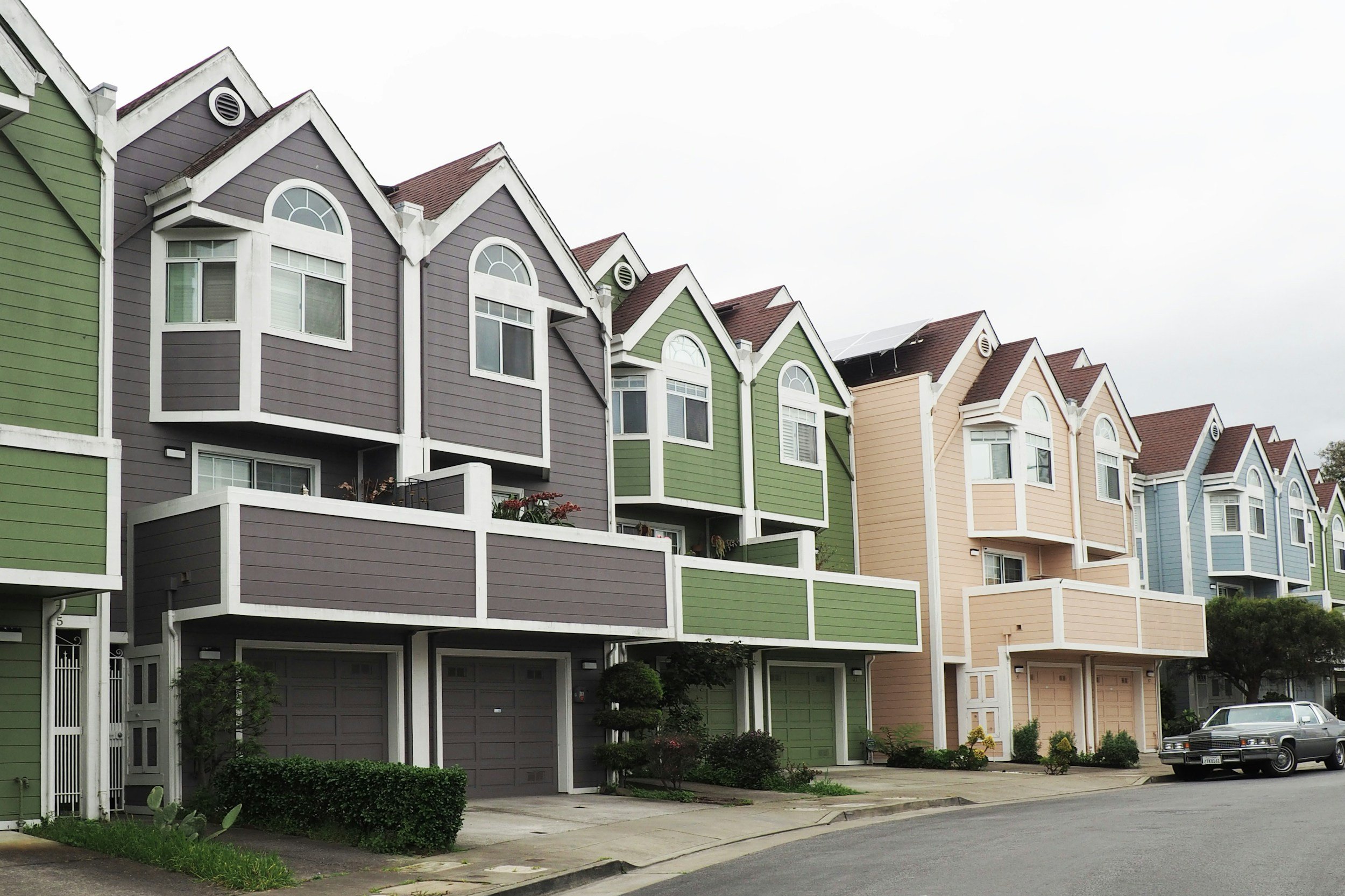 Colorful townhouses with white trim, located along a curved street with parked cars, under an overcast sky.
