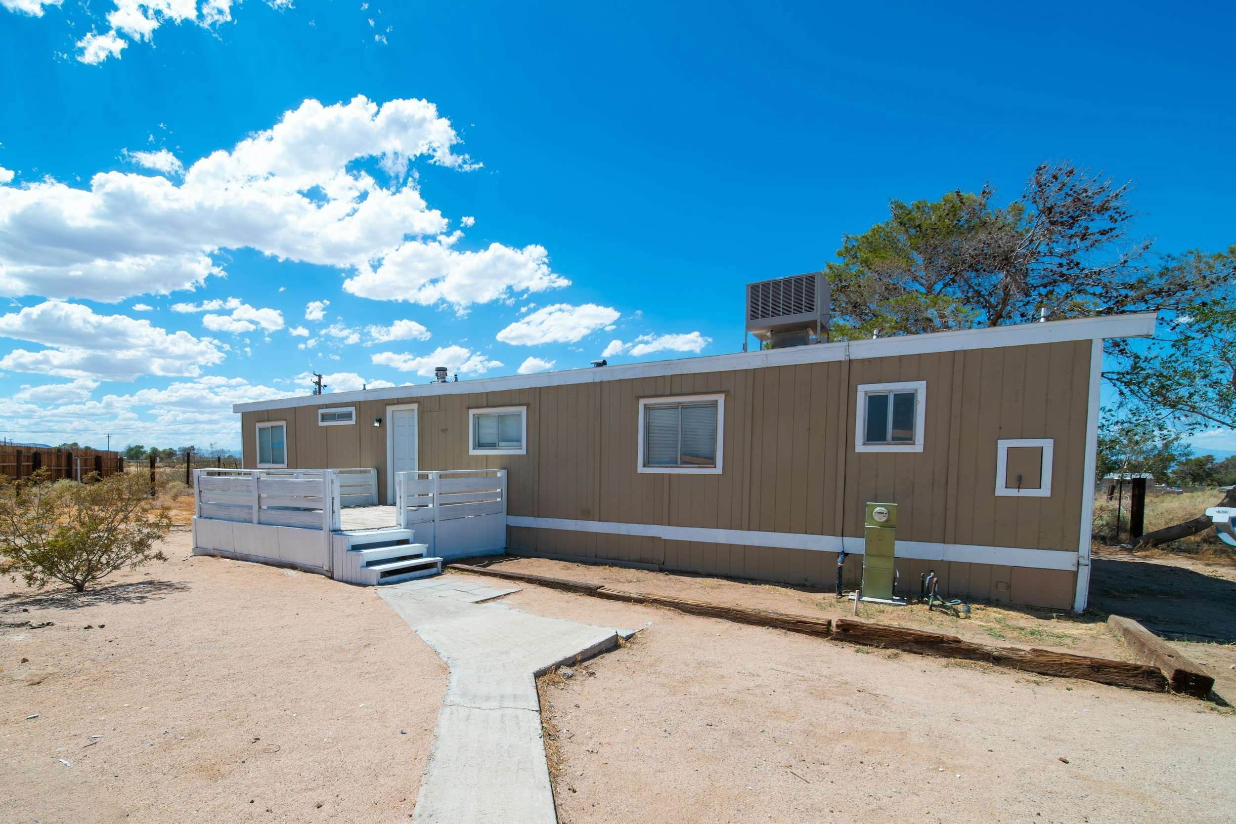 A single-story beige manufactured home with white trim, situated in a desert landscape with sparse shrubbery, under a bright blue sky with scattered clouds. The house has a small front porch with steps, two windows, and an air conditioning unit on the roof.