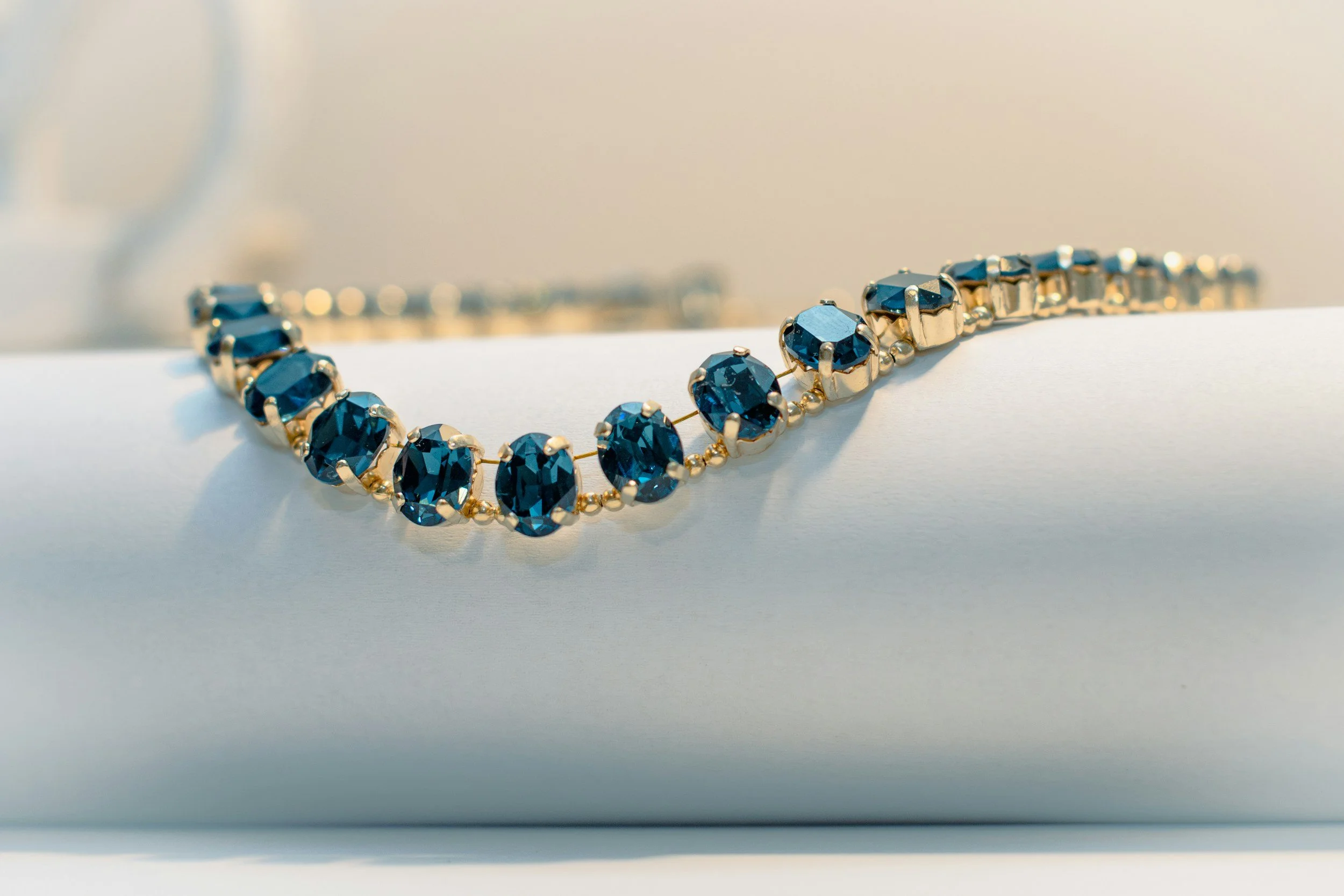 A gold bracelet with oval-cut green gemstones is displayed on a white cylindrical surface.