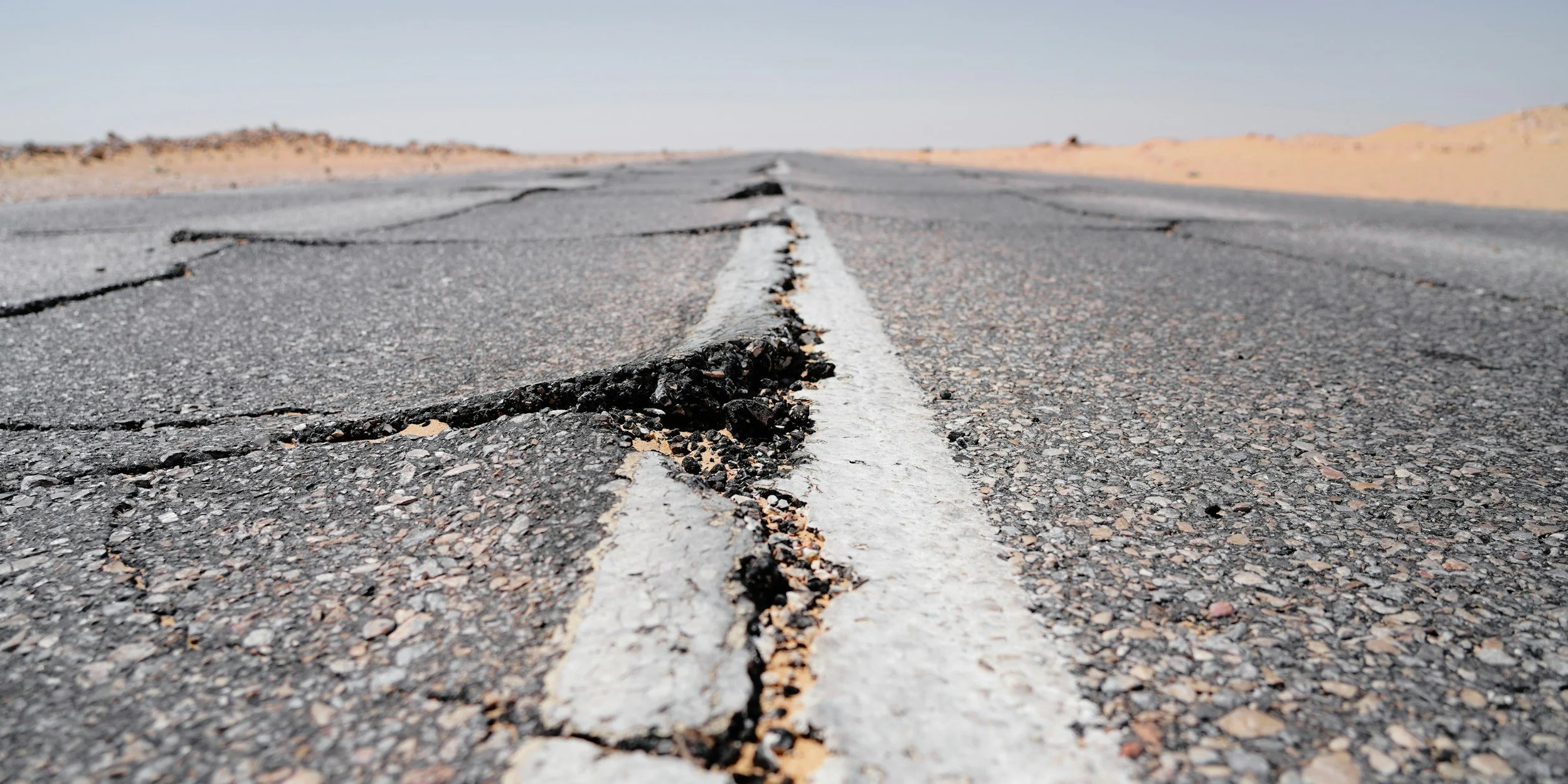Close-up of a cracked and damaged asphalt road in a desert landscape, with distant sand dunes under a clear sky.