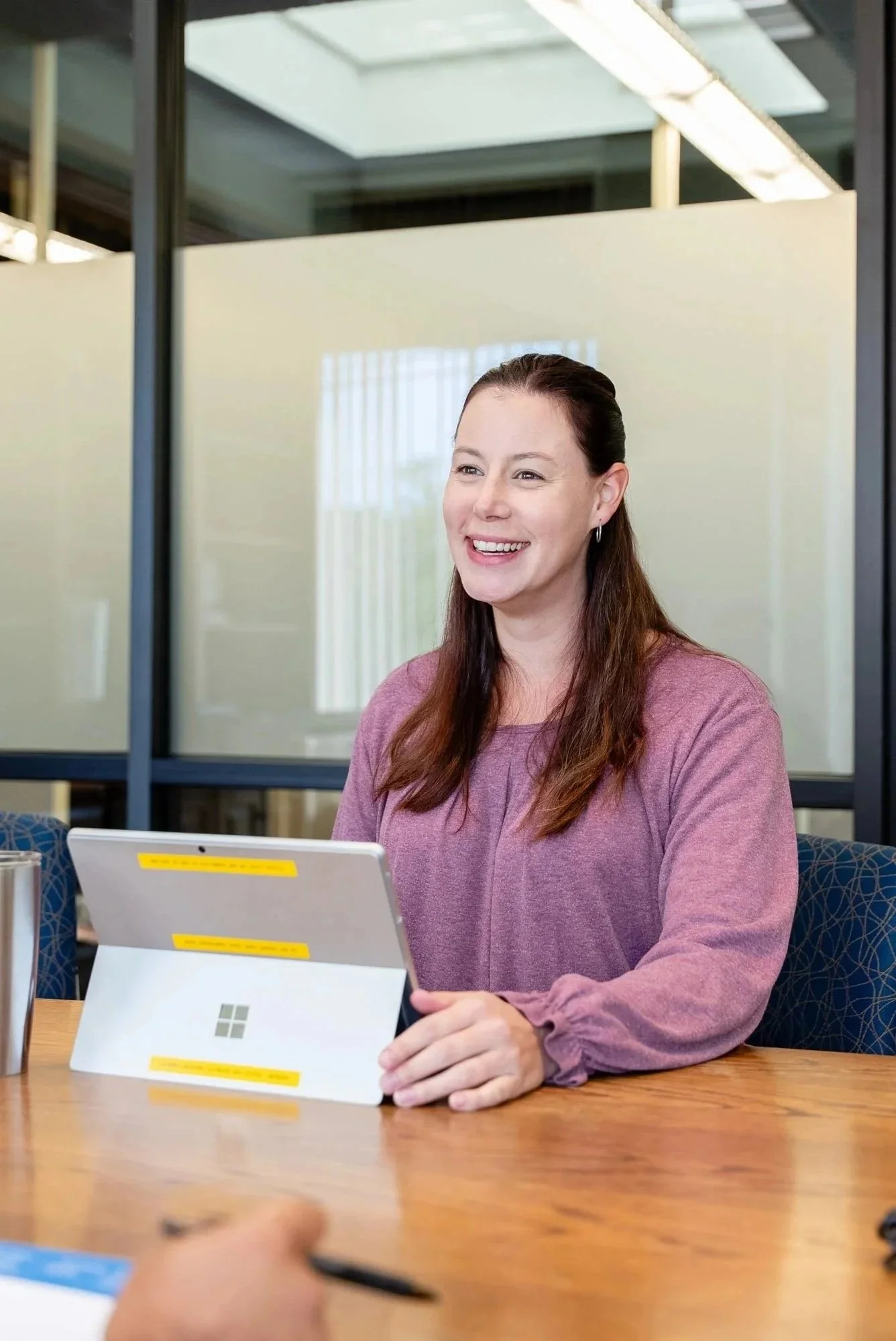 A woman with long brown hair smiling while sitting at a table with a tablet in front of her, in a modern office setting.