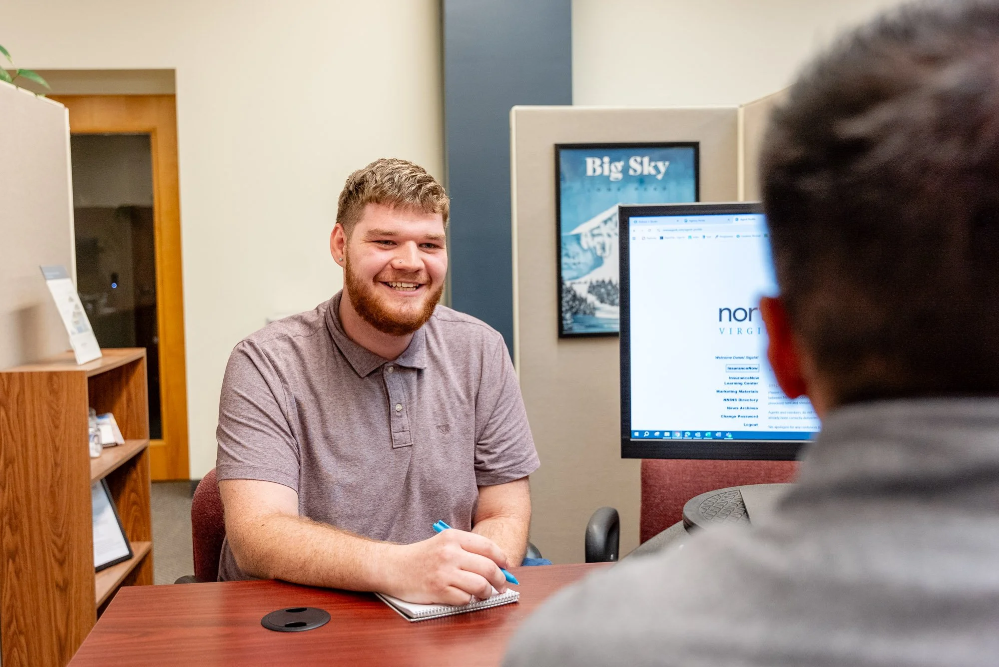 A young man with a beard and a grey polo shirt sitting at a desk, smiling during a conversation with a person whose back is to the camera, in an office setting with a computer monitor, a framed picture of Big Sky, and a wooden bookshelf.