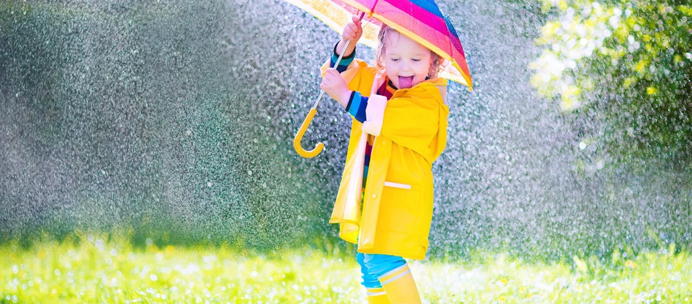 A young girl in yellow raincoat and rain boots playing in the rain with a colorful umbrella on a bright, rainy day.