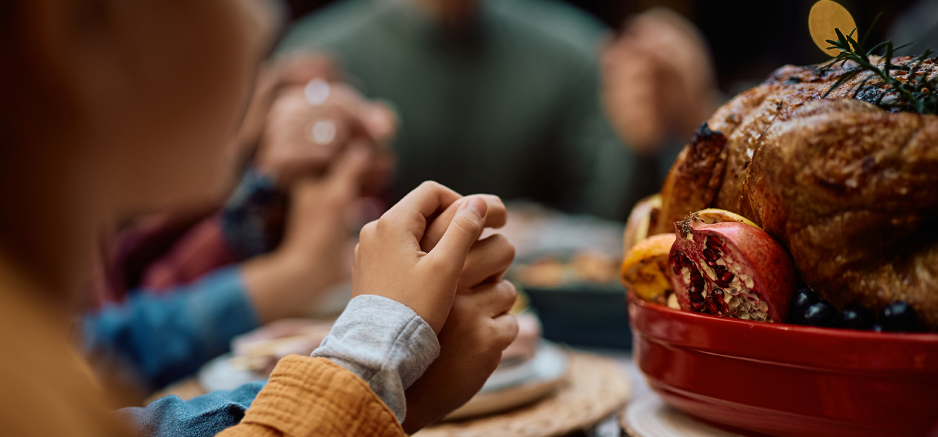 People holding hands at a dinner table with a roasted turkey in the foreground.