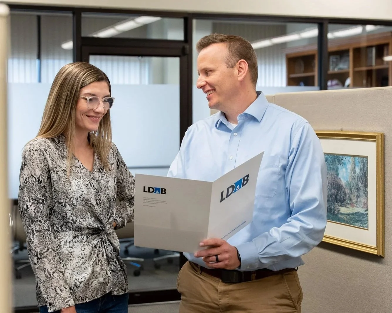 Two colleagues, a woman with long blonde hair and glasses wearing a patterned blouse, and a man with short hair in a blue shirt and khakis, stand in an office discussing a document labeled 'LD&B.' They are smiling and looking at each other, with office cubicles and framed artwork in the background.