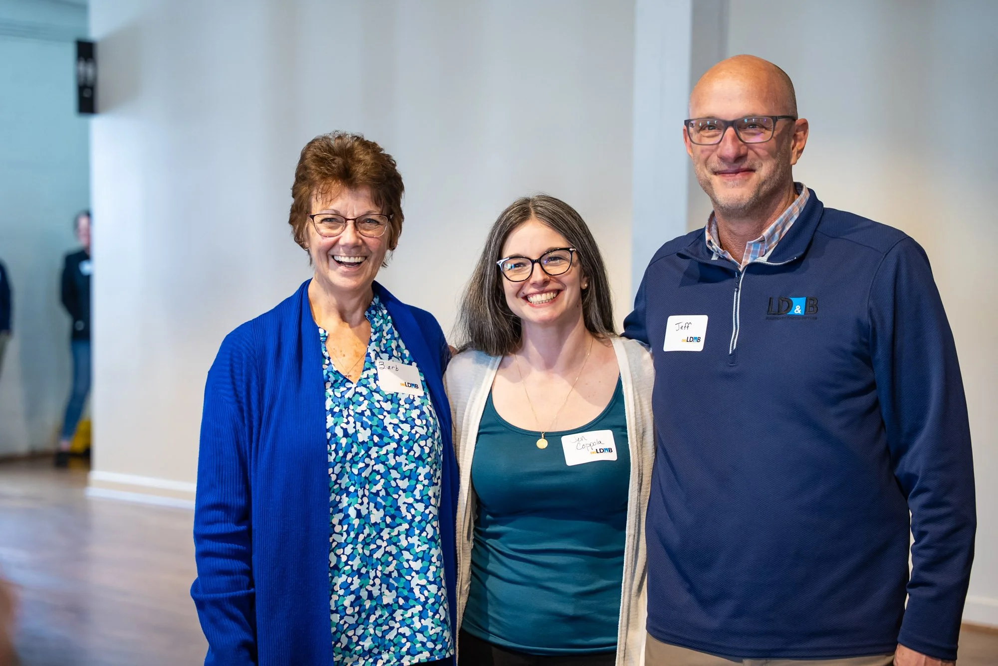 Three smiling people standing together at an indoor event, wearing name tags. The woman on the left has short brown hair, glasses, and is wearing a blue cardigan over a floral shirt. The woman in the middle has long gray hair, glasses, and is wearing a teal shirt with a beige cardigan. The man on the right has a shaved head, glasses, and is wearing a navy blue jacket with a name tag that says Jeff.