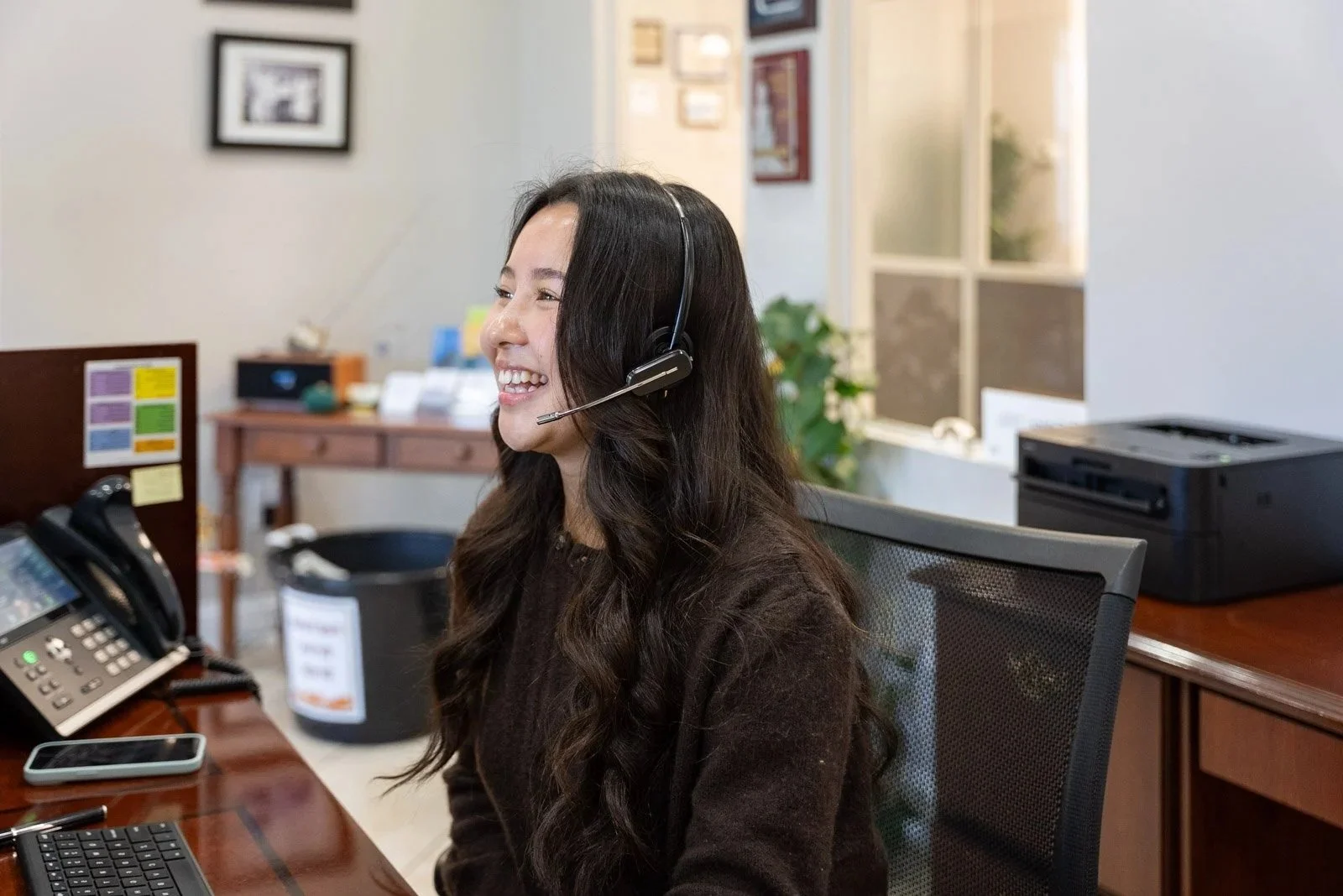 A woman with long dark hair wearing a headset, sitting at a desk in an office, smiling and looking to the left.
