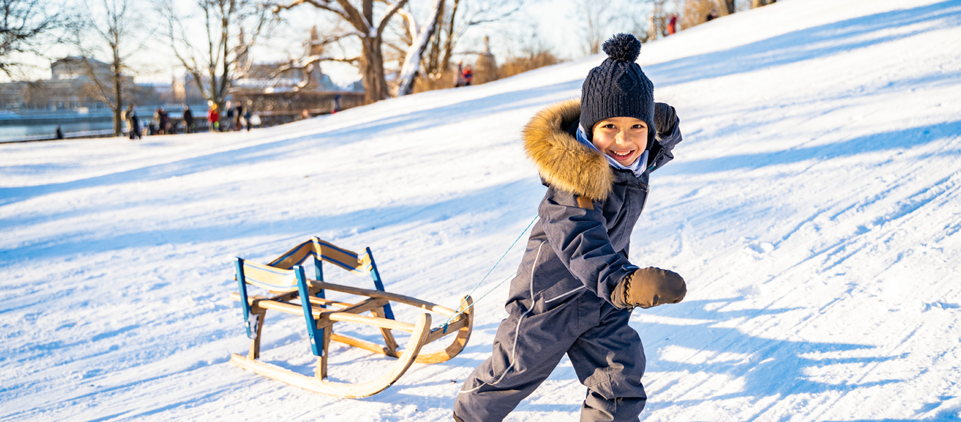 A young boy in winter clothing, including a beanie, gloves, and a coat with a fur-lined hood, smiling while pulling a wooden sled up a snowy hill during daytime.
