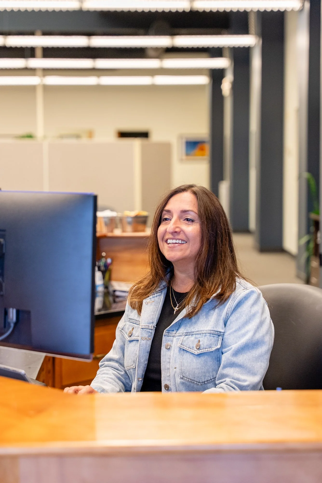 A woman with brown hair smiling at her computer in an office setting, wearing a denim jacket and black shirt.