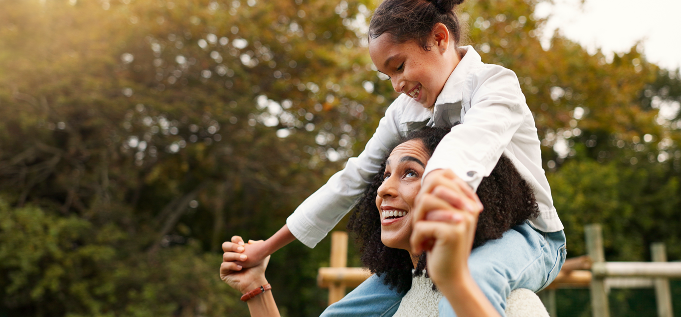 A woman with curly hair giving a piggyback ride to a young girl with hair tied back. They are outdoors in a park with autumn-colored trees.