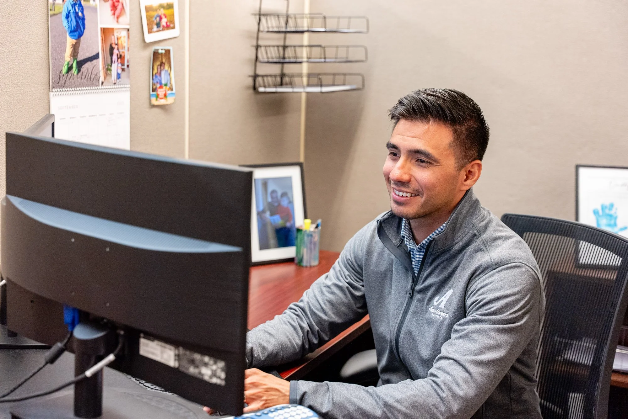 A man with short dark hair smiling at his computer monitor in an office. He is wearing a gray jacket over a patterned shirt. There is a computer desktop, office supplies, and a monitor on his desk, with photos on the wall behind him.