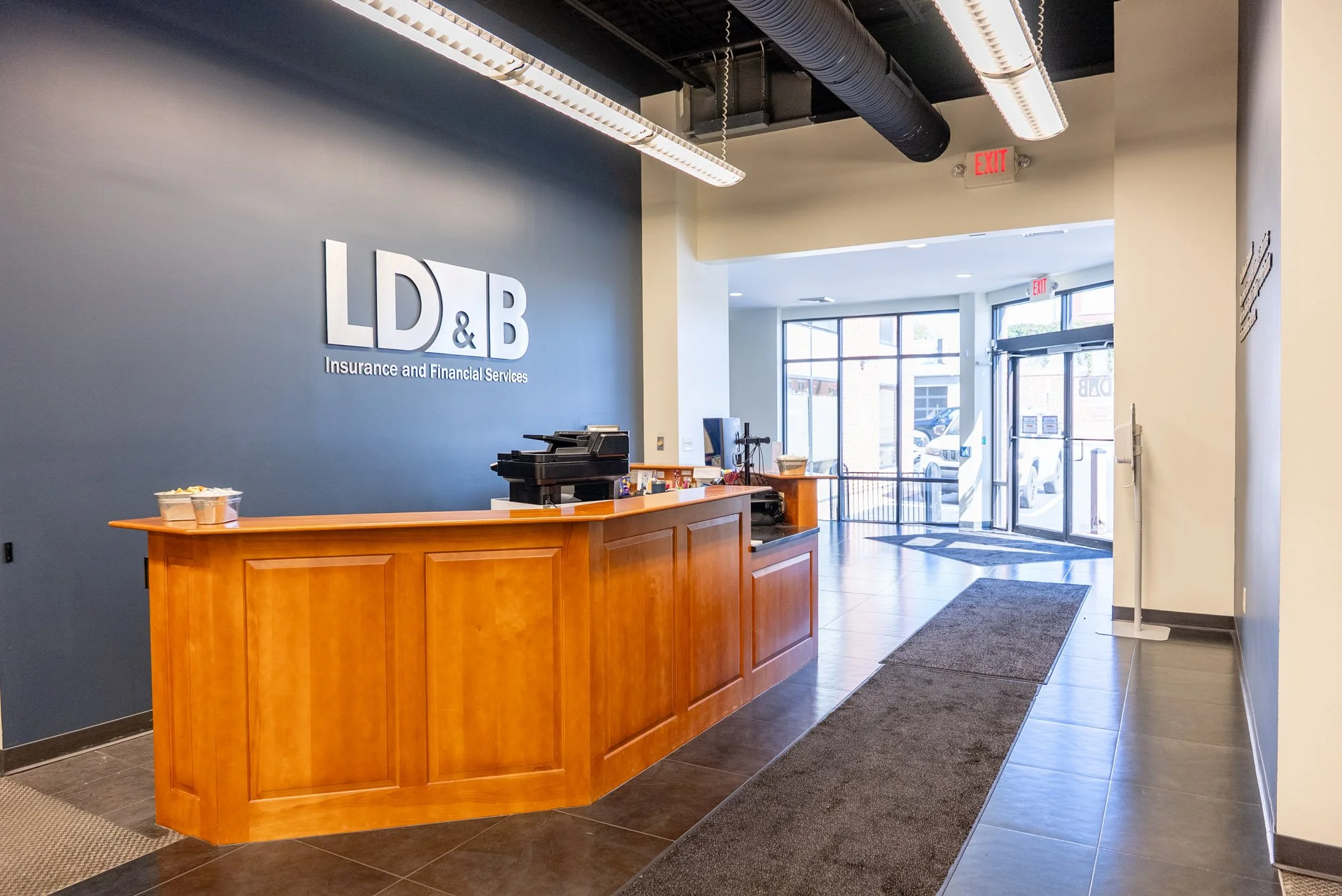 Reception area of the LD&B office with a blue wall displaying a sign that says "LD&B Insurance and Financial Services", wooden counter with office equipment, glass entrance doors, and large windows letting in natural light.