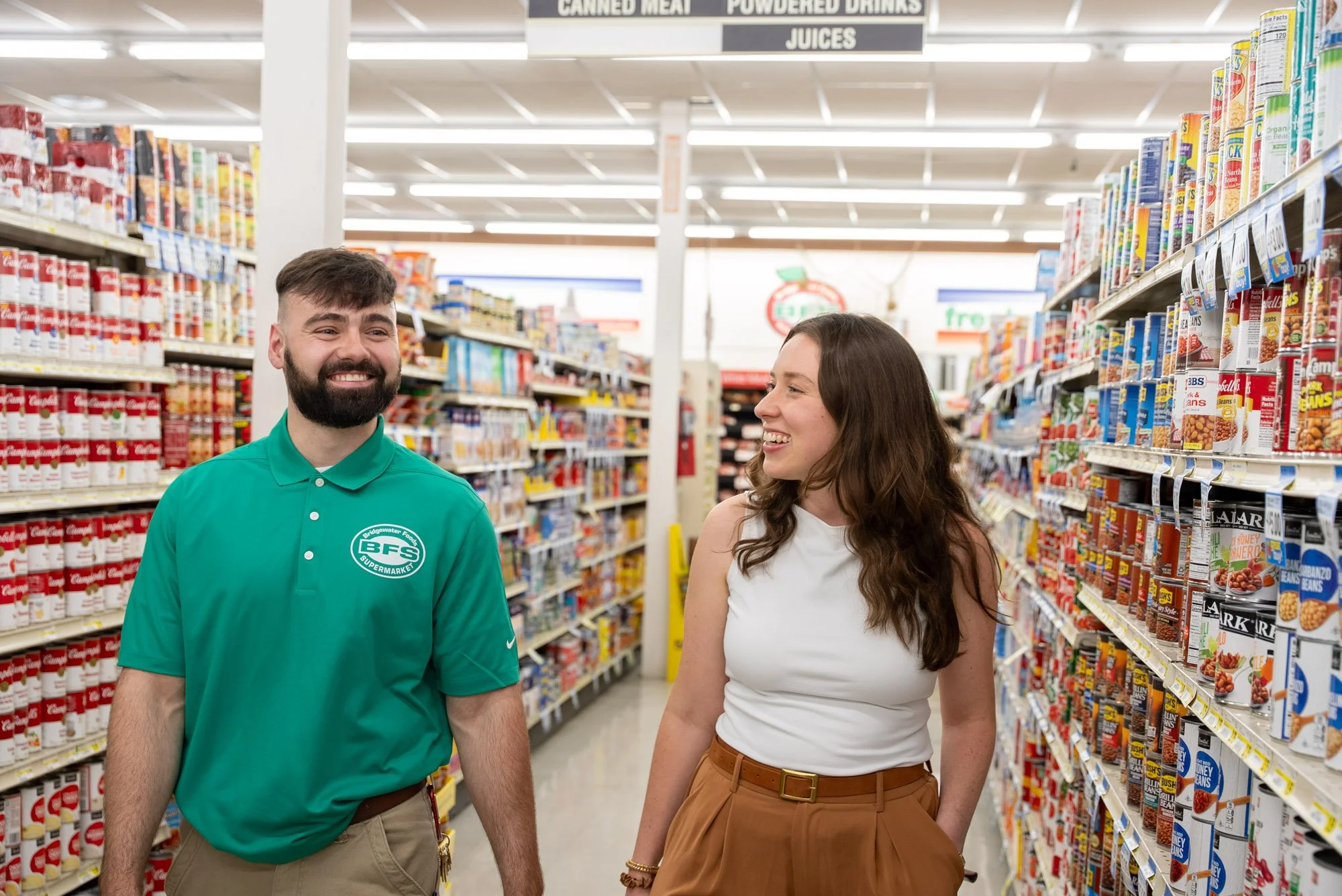 A man and woman smiling and talking in a grocery store aisle filled with canned goods and other food products.