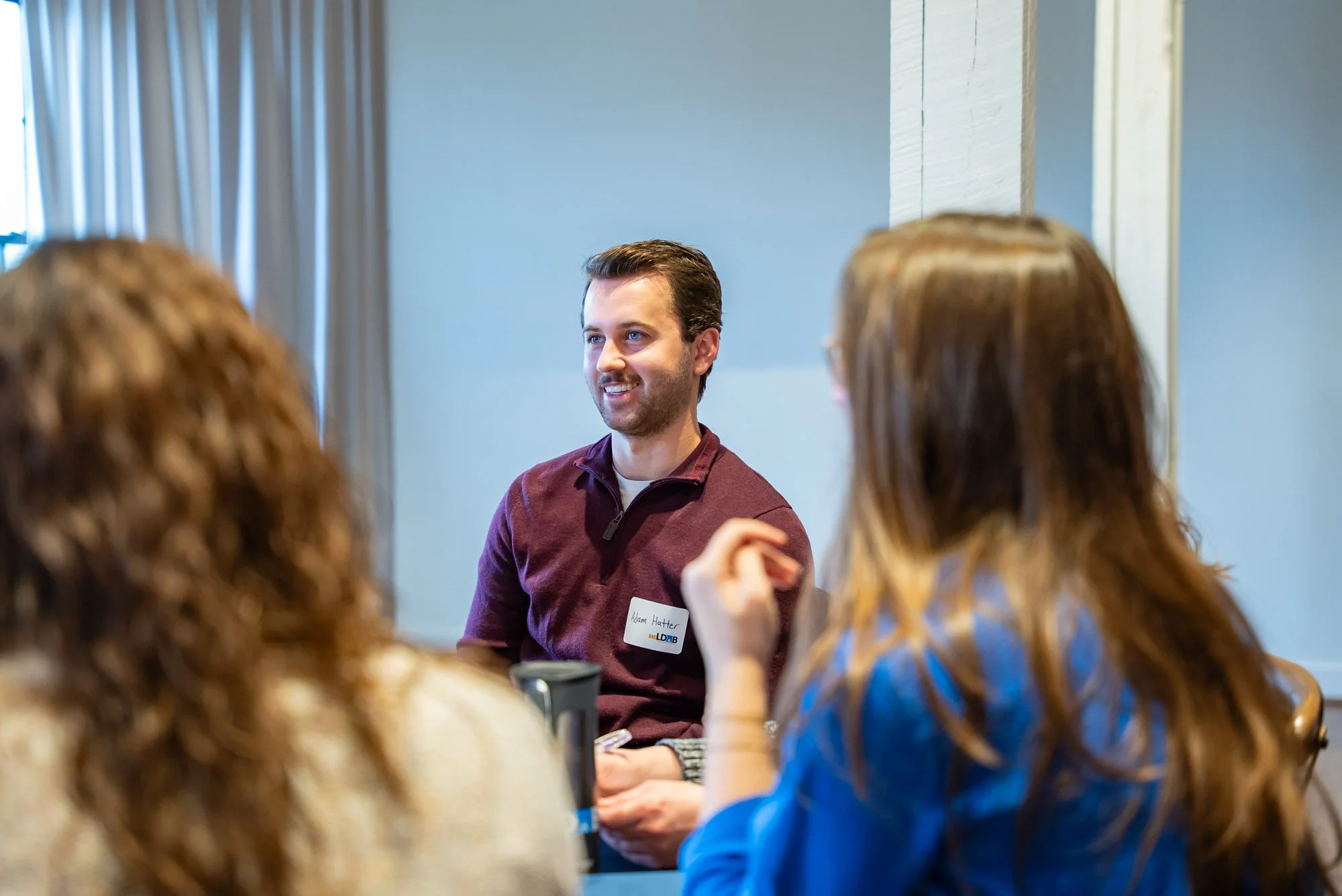 A man with a name tag speaking to two women during a meeting in a conference room.