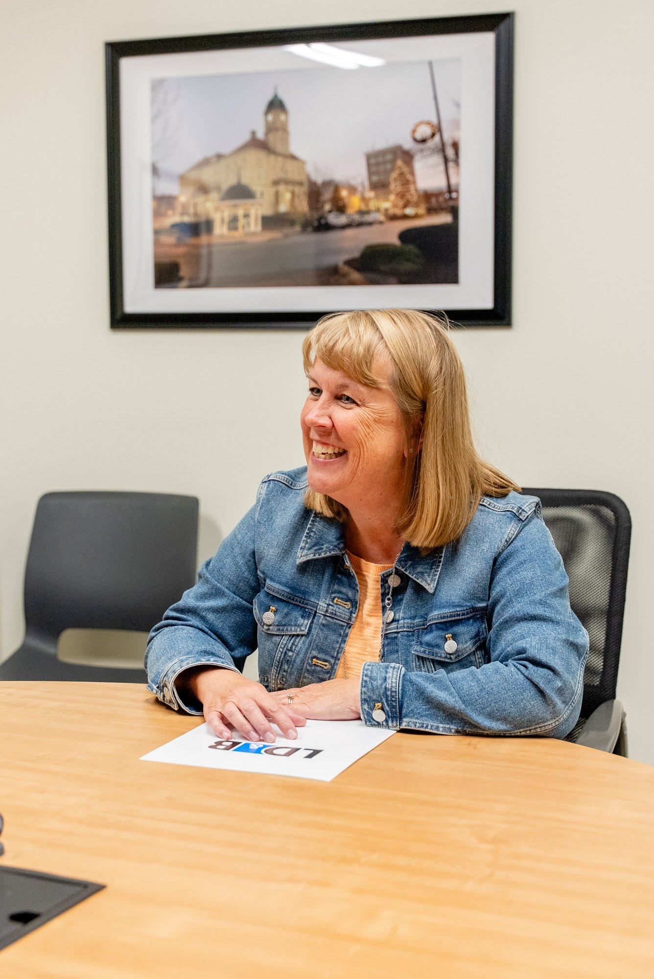 A woman with blonde hair wearing a denim jacket, smiling and sitting at a wooden table in a meeting room.