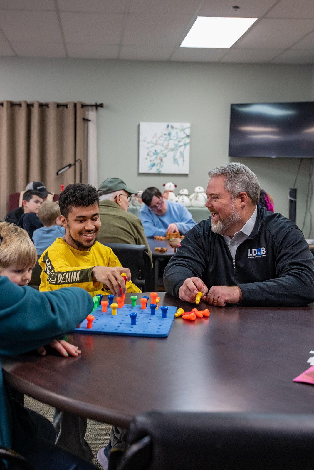 People playing Connect Four game at a table, smiling and enjoying their time, in a room with a television and decorative artwork on the wall.