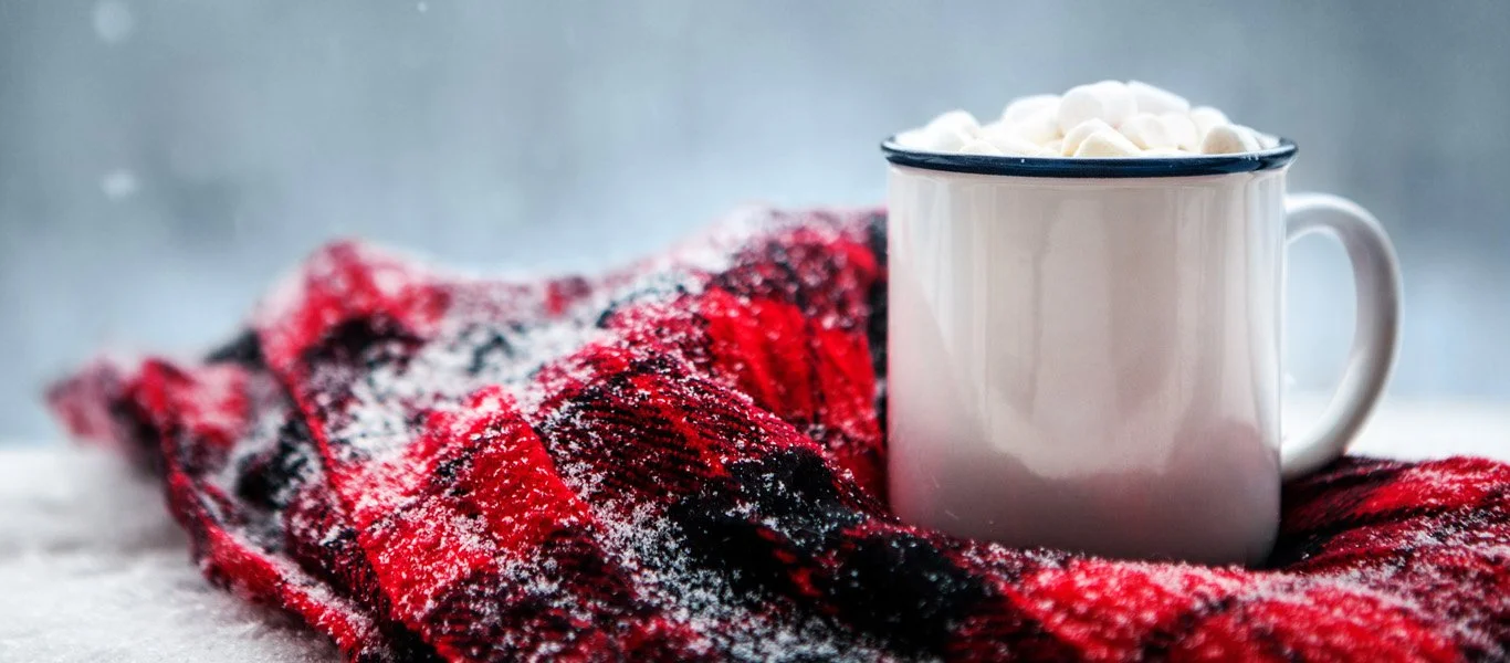 A white mug filled with marshmallows resting on a red and black plaid blanket with snow dusting.