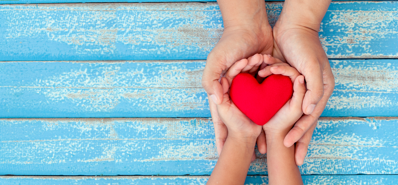 Adult hands and child's hands holding a red heart over a distressed blue wooden surface.