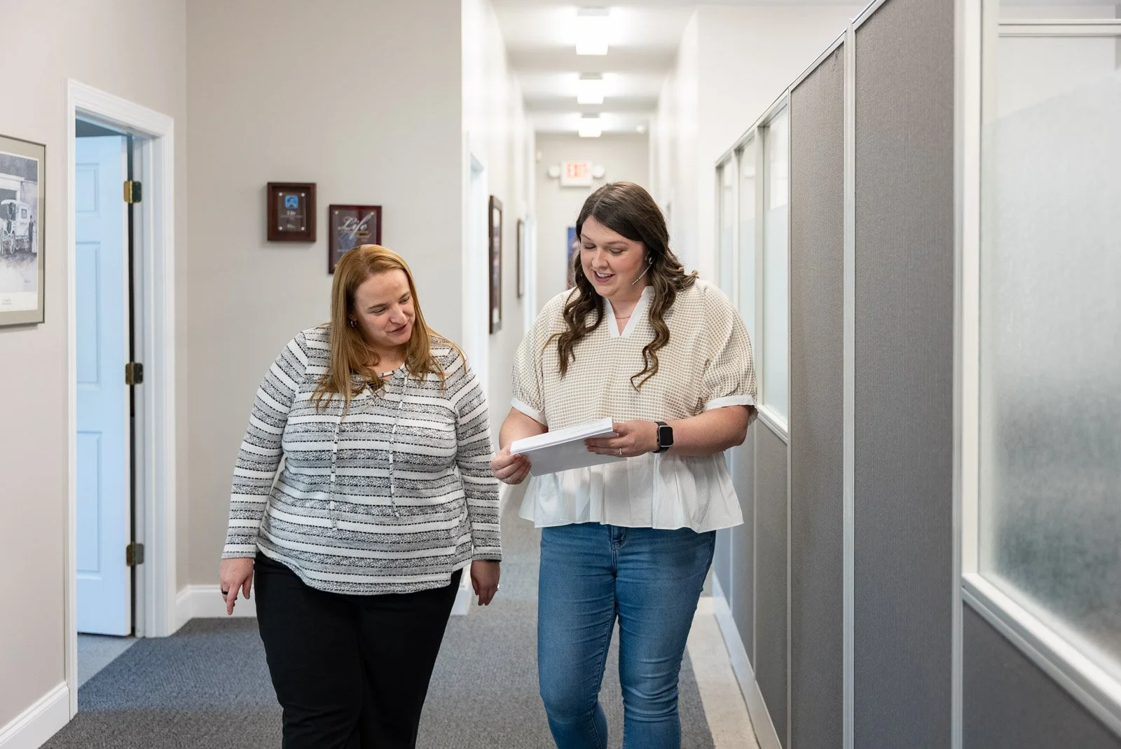 Two women walking and talking in an office hallway, one is holding papers and smiling, the other is listening and looking at the papers.