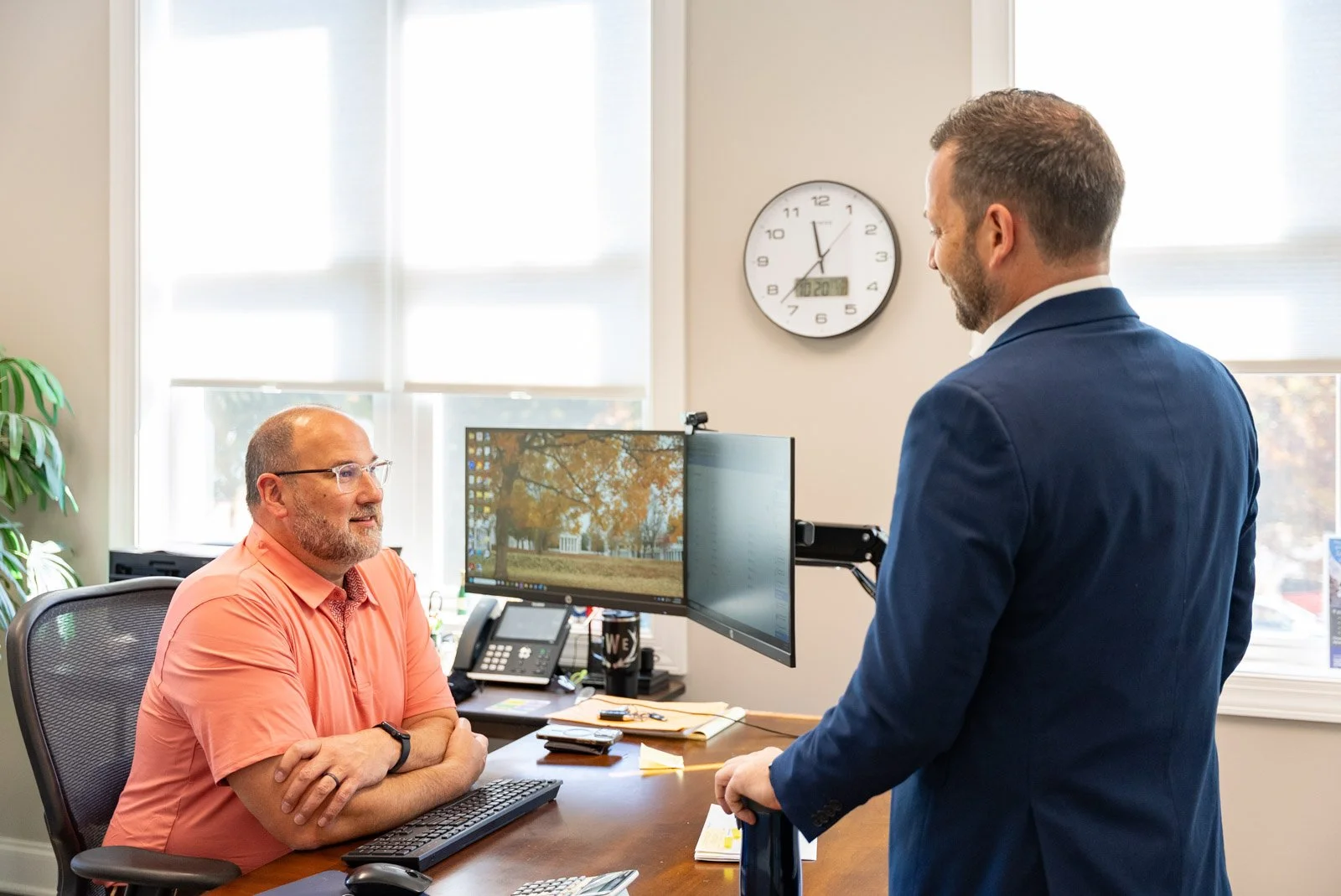 Two men in an office engaged in conversation, one seated at a desk with a computer and the other standing in front of him. The seated man wears glasses and a coral shirt, smiling, and the standing man wears a blue suit.